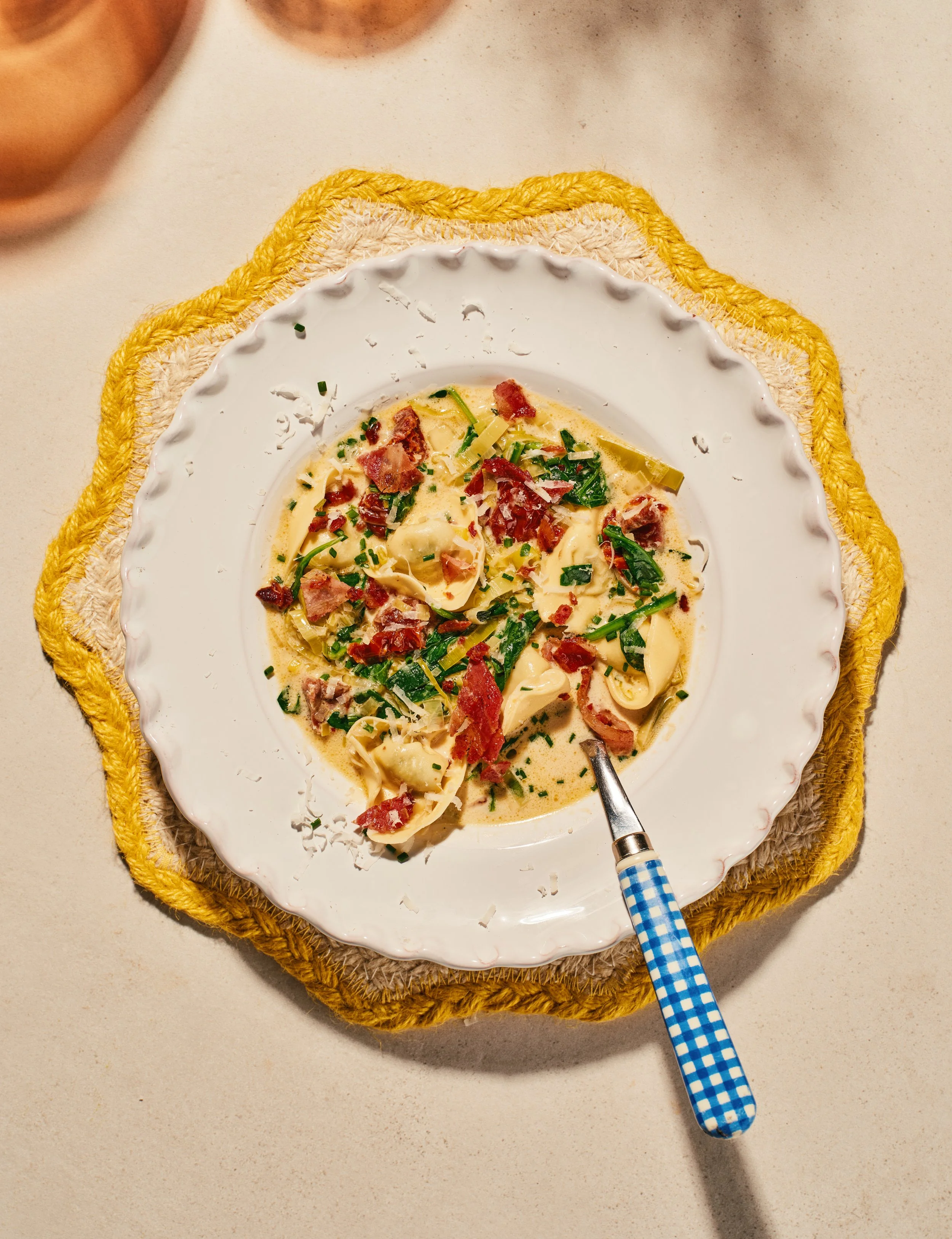 A bowl of creamy pasta with bacon and green herbs on a white plate, placed on a woven placemat on a white table, with a blue checkered spoon inside the bowl.