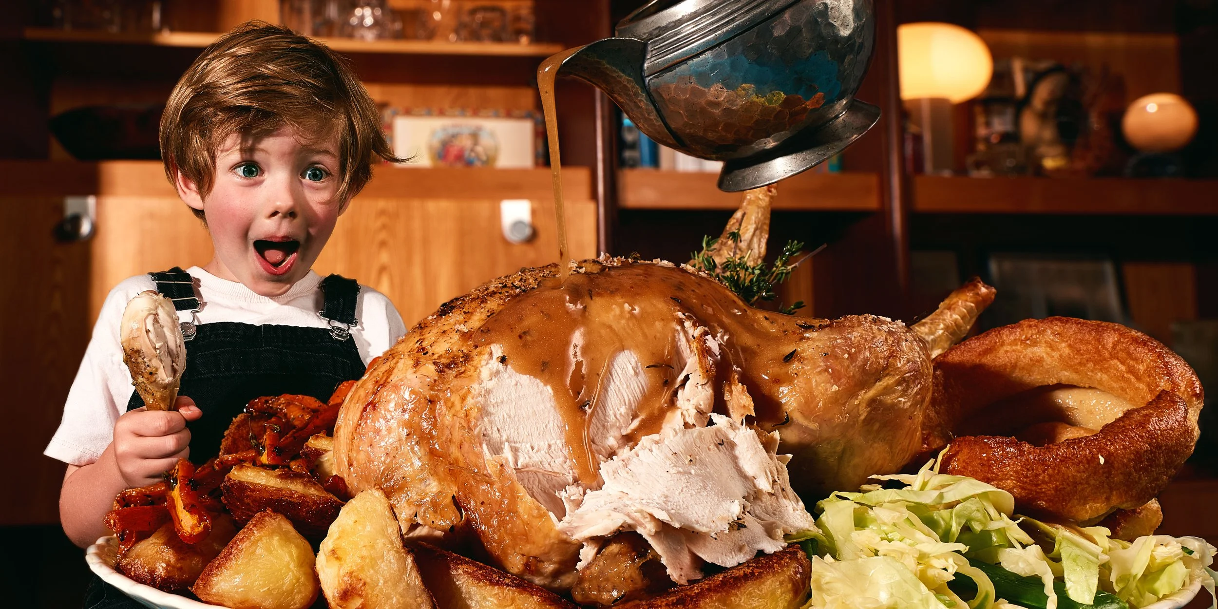 A young child with excited expression holding a drumstick near a large roasted chicken dinner with potatoes and vegetables in a cozy kitchen.