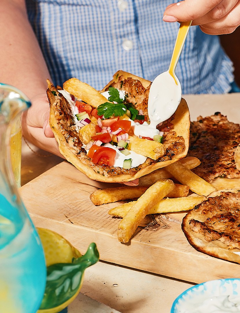 Person holding a taco filled with grilled meat, French fries, chopped tomatoes, cucumbers, red onions, cilantro, and sour cream with French fries on a wooden serving board along with grilled meat and a blue drink with lemon in a mug.