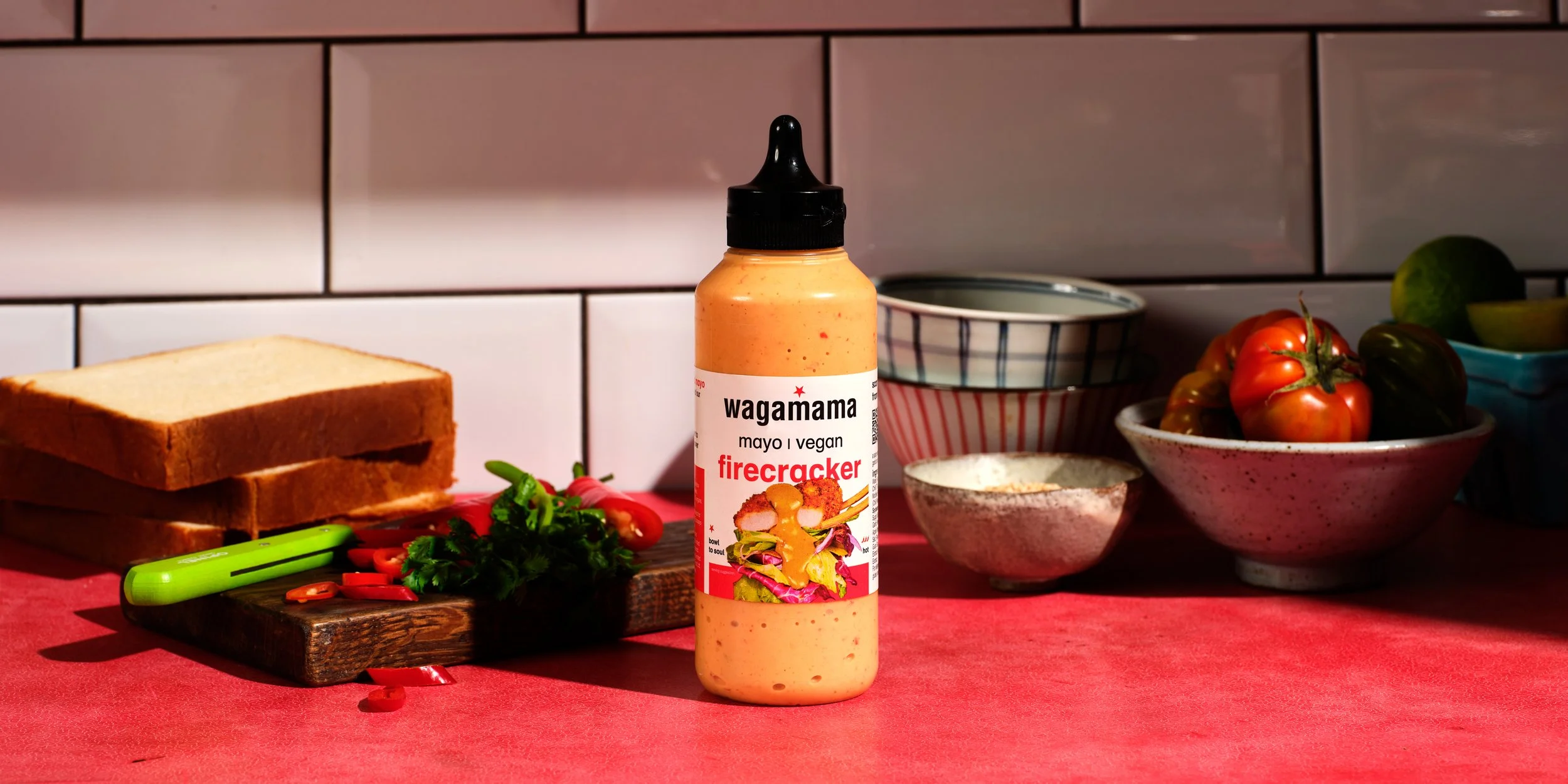 A bottle of Wagamama firecracker vegan mayo sauce on a red table with sliced bread, cherry tomatoes, and various bowls of vegetables in the background.