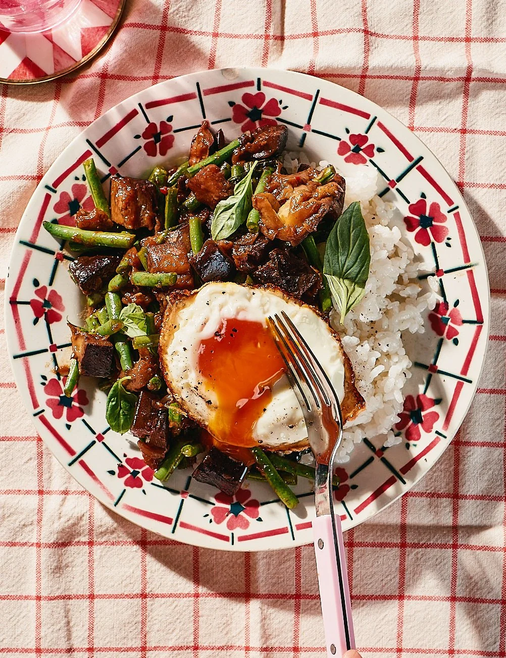 A plate of Asian-inspired food with rice, green beans, eggplant, sliced meat, fresh basil, and a soft-boiled egg with runny yolk, on a red and white checkered tablecloth.