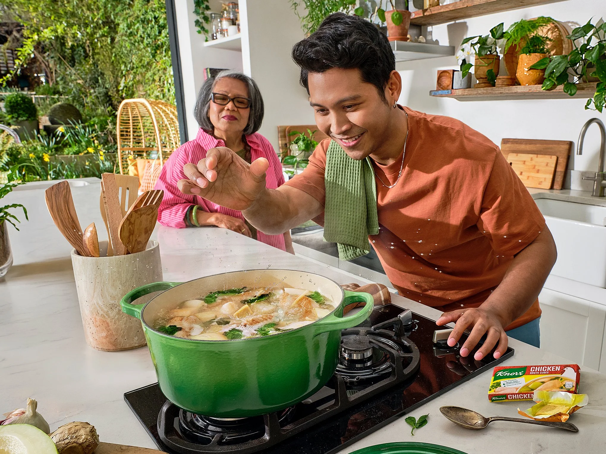 A young man cooking soup on a stove in a kitchen with an older woman nearby. The kitchen has a window showing outdoor greenery, and there are shelves with potted plants and kitchen items. The table has garlic, a spoon, and packages of Knorr chicken b