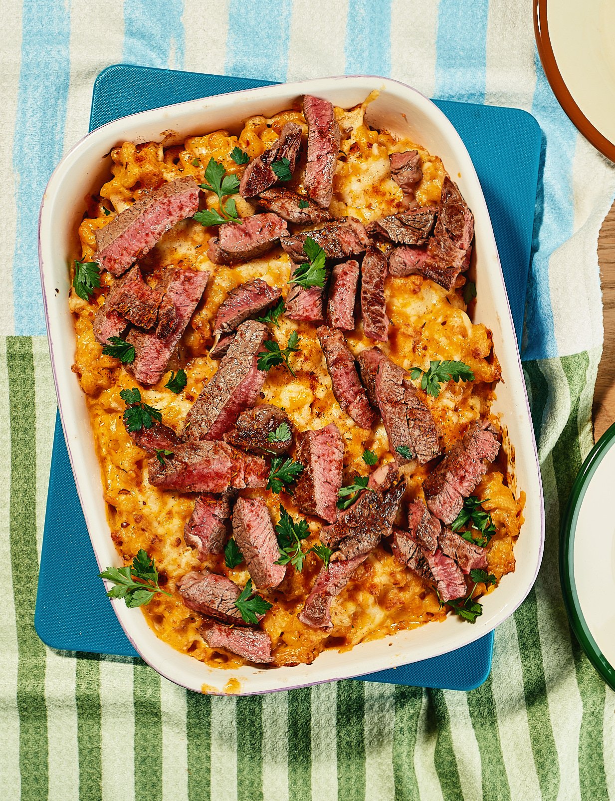 Baked macaroni and cheese topped with sliced cooked steak and garnished with fresh parsley, in a white dish on a blue placemat.