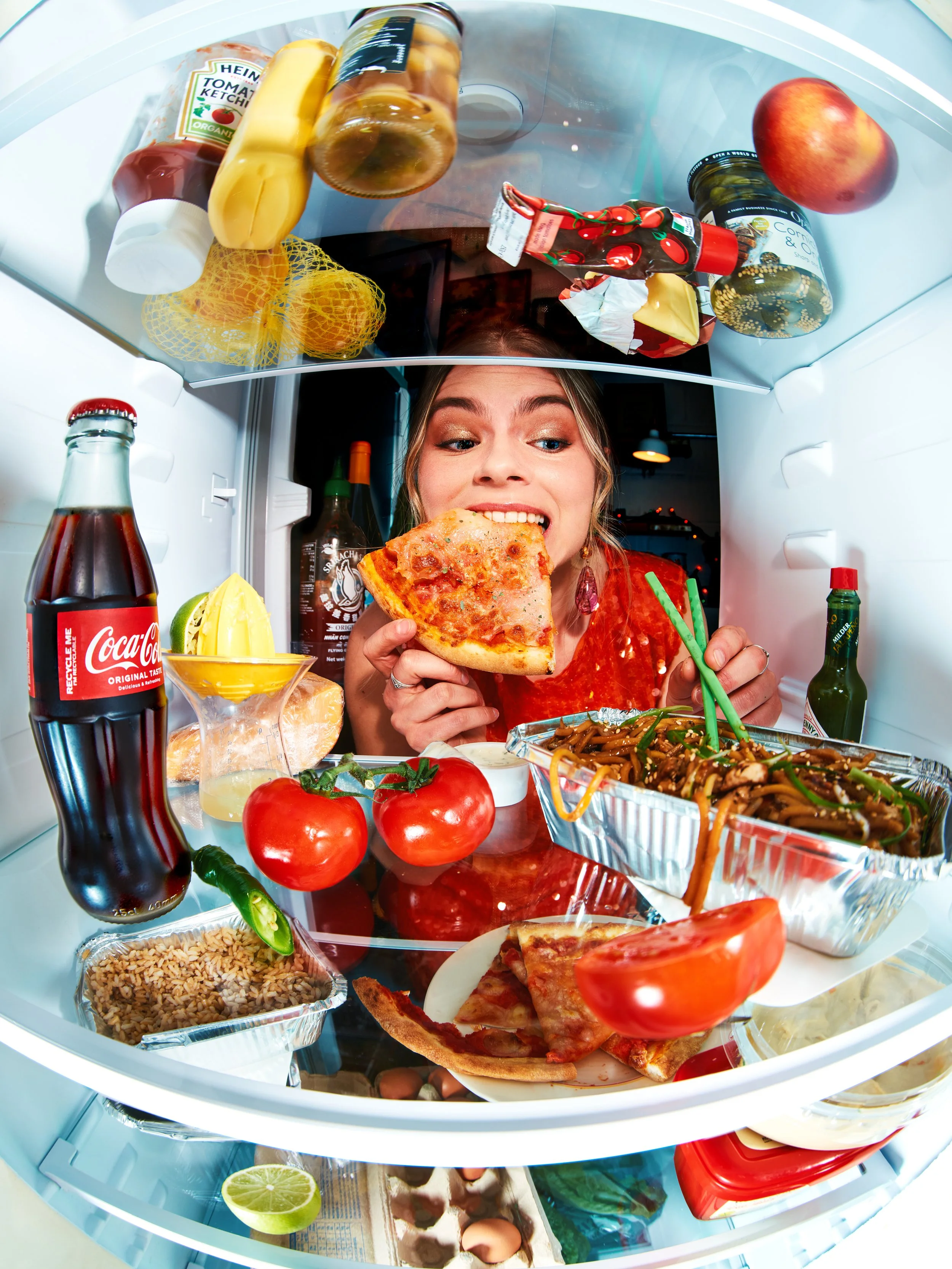 Inside a refrigerator, a woman is smiling and holding a slice of pizza, surrounded by various foods such as tomatoes, lime, soy sauce, noodles, and condiments.