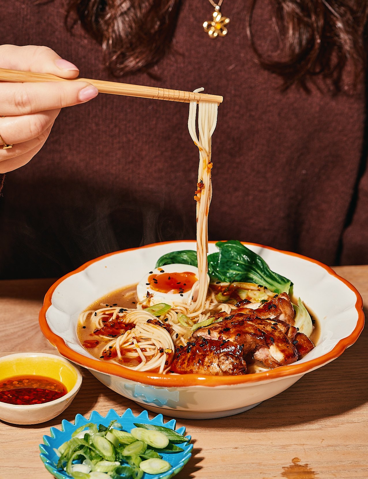 A person holding chopsticks lifting noodles from a bowl of ramen with sliced chicken, boiled egg, greens, and black sesame seeds, with dipping sauces and sliced green onions on the side.