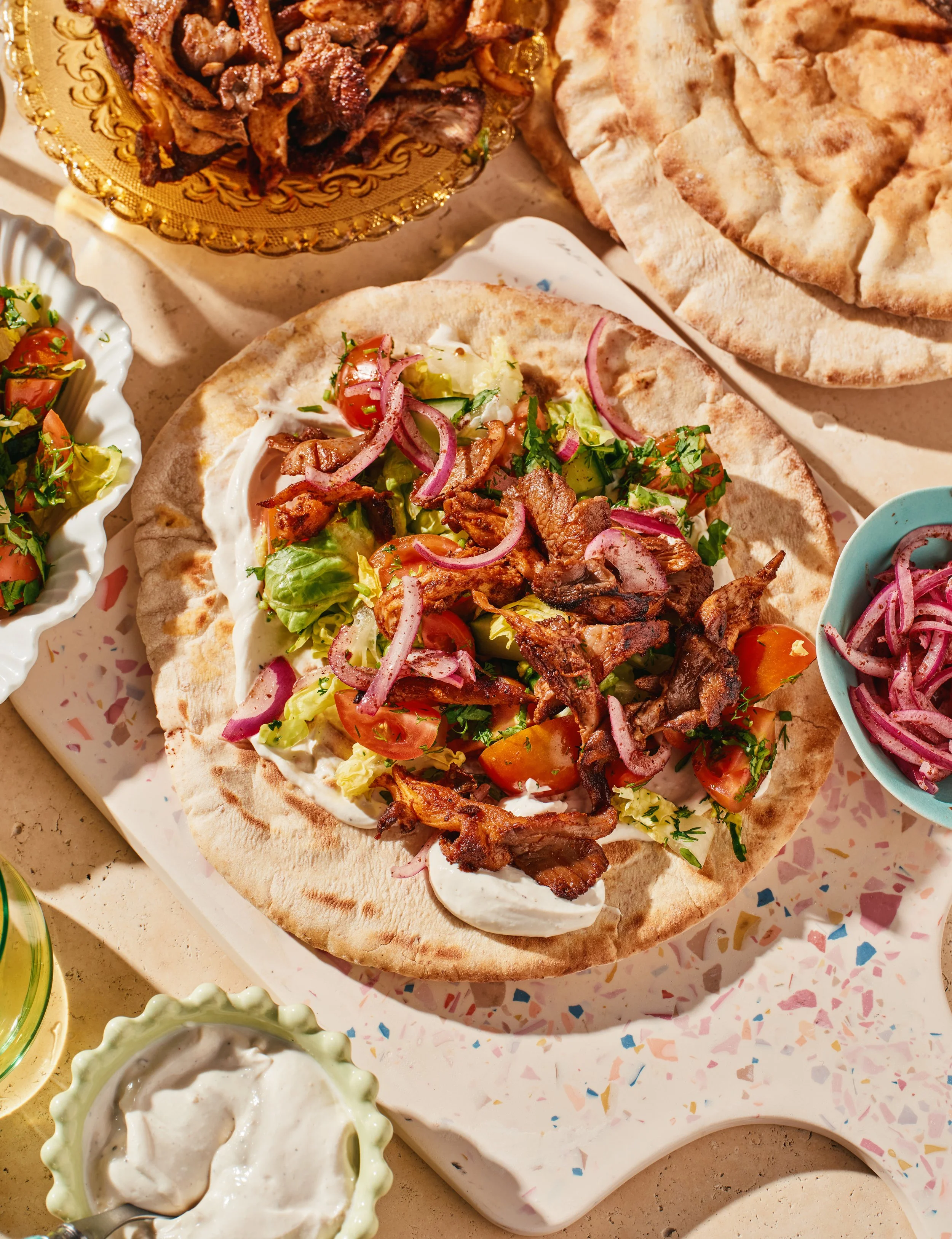 A plate with a naan bread topped with grilled meats, fresh vegetables, and herbs, alongside small bowls of yogurt-based sauces and salads.