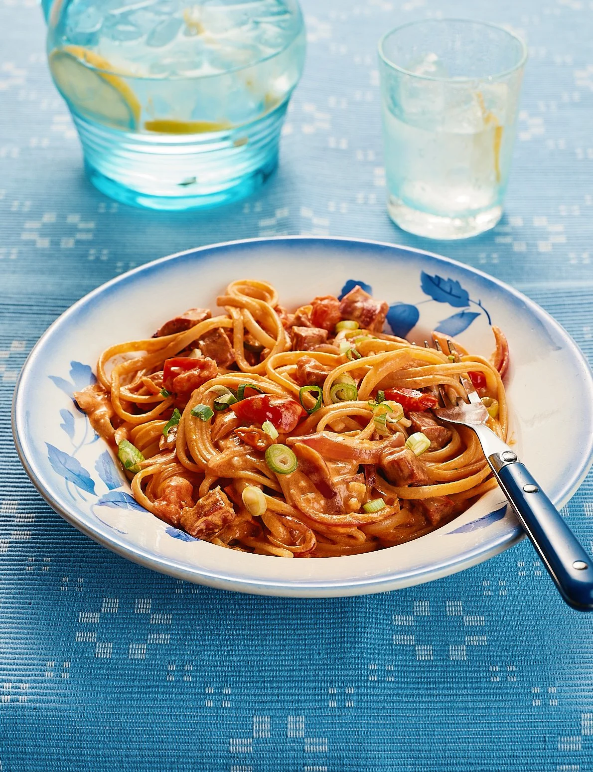 Plate of pasta with meat, cherry tomatoes, and green onions on a blue tablecloth, with a fork in the pasta, and a glass pitcher and glass of water in the background.