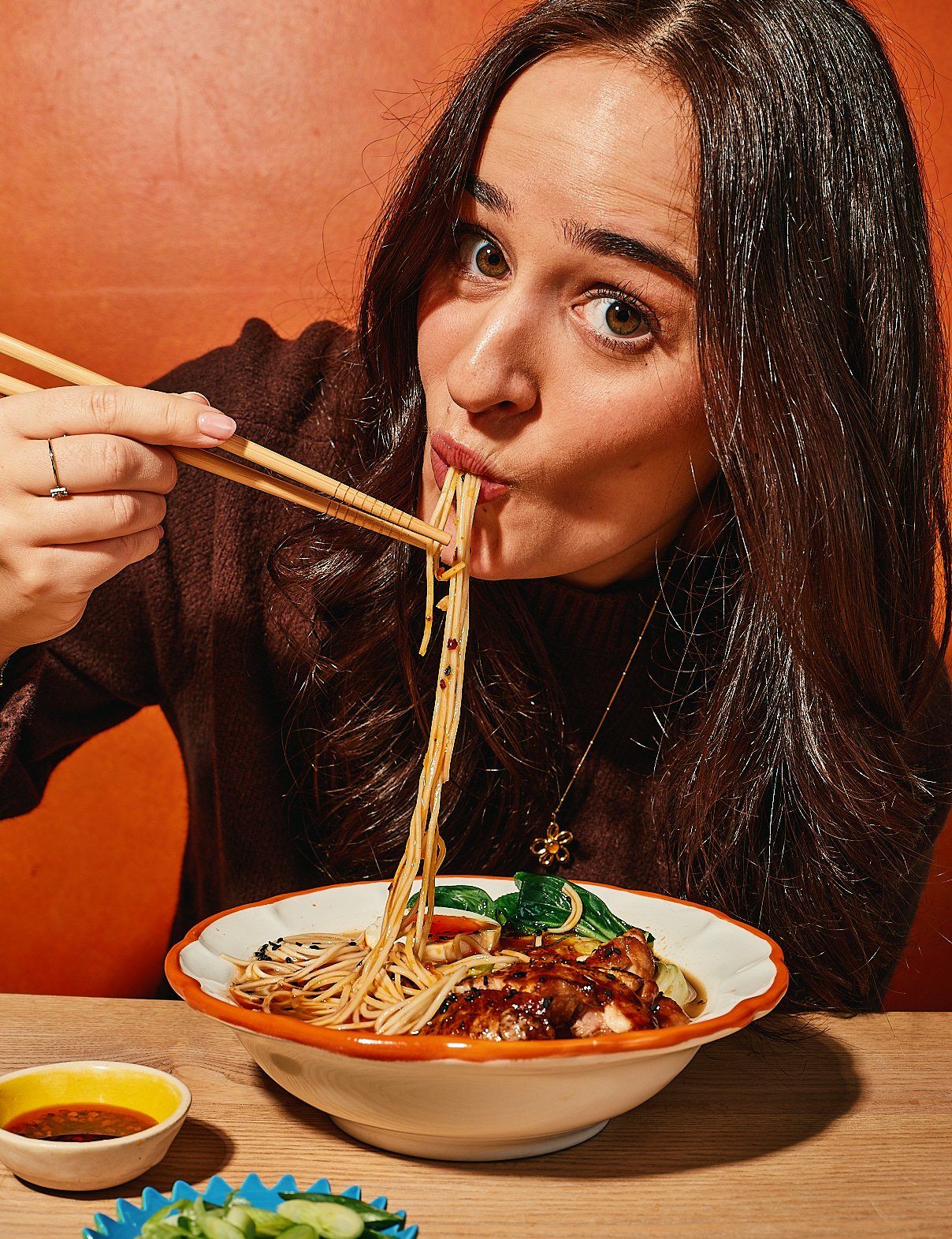 Woman with long dark hair eating Asian noodles with chopsticks at a restaurant, with a bowl of noodles and vegetables in front of her