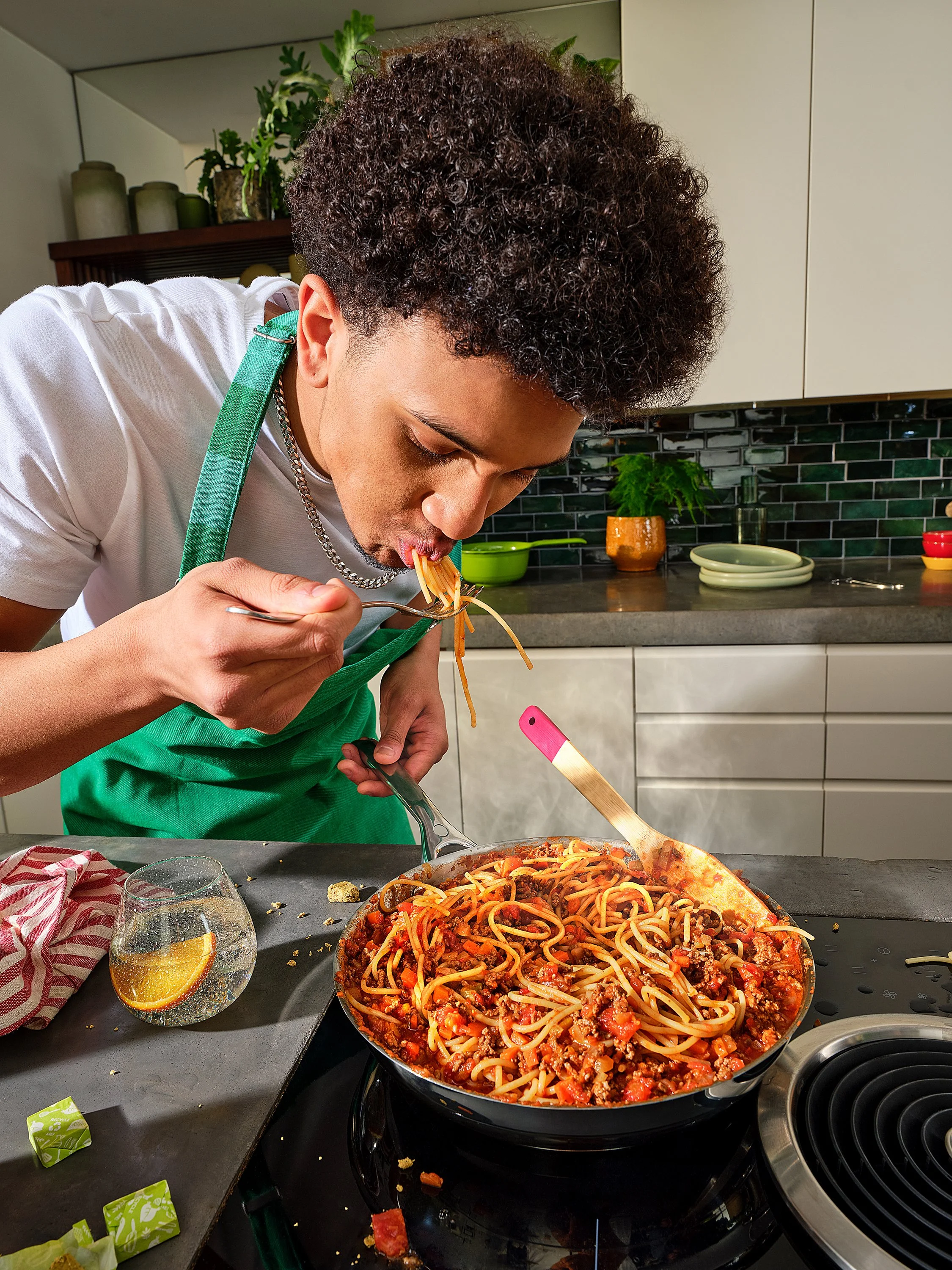A person with curly hair and wearing a white shirt and green apron is eating spaghetti with meat sauce from a fork in a kitchen. There is a pan of spaghetti on the stove, a glass of water with lemon, a red and white checkered cloth, and some small gr
