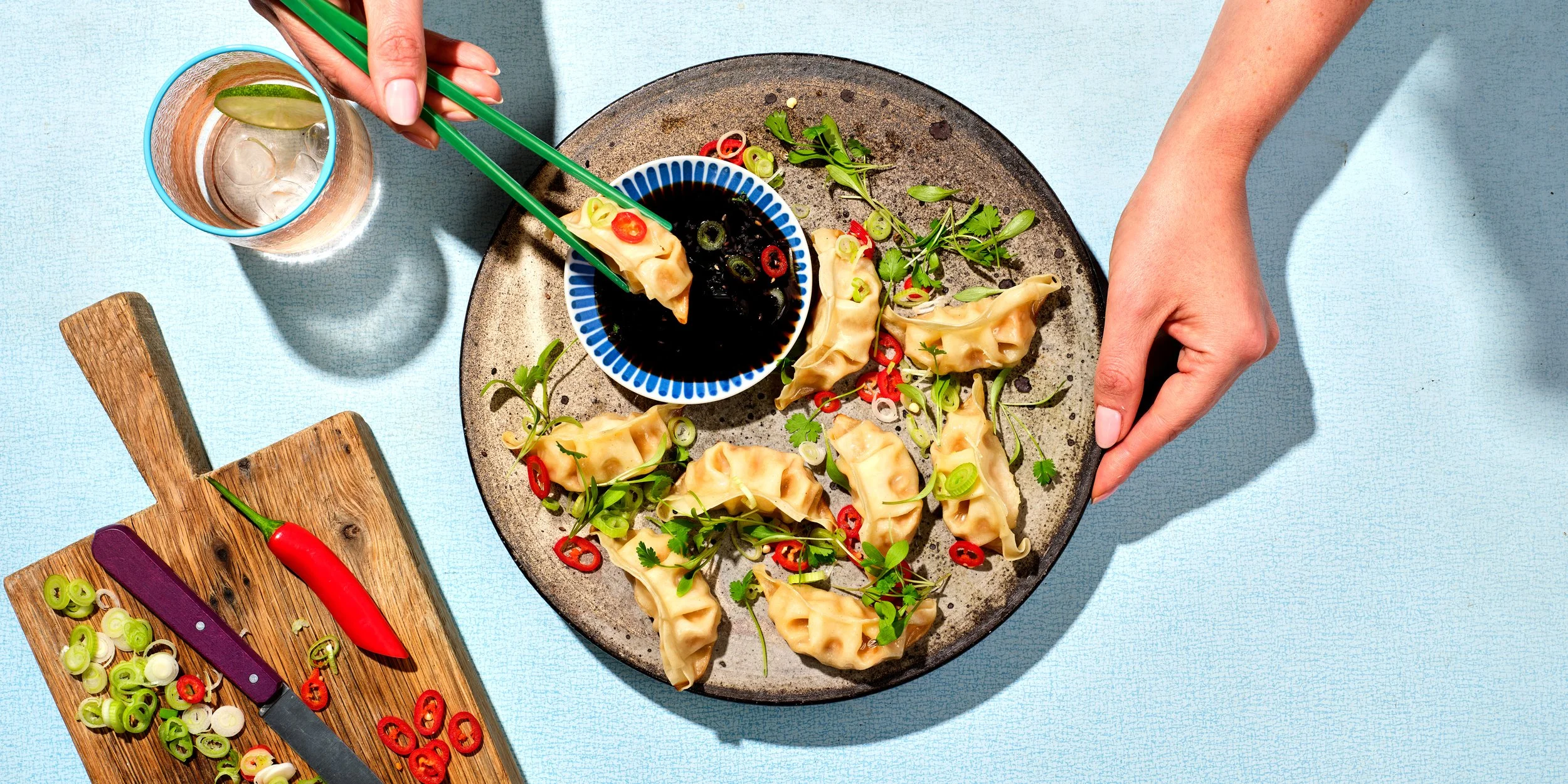 Person using green chopsticks to pick up a dumpling from a plate with more dumplings, sliced green onions, and red chili slices, on a blue surface. A glass of drink with ice and lime is nearby. A wooden cutting board with sliced green onions, red chi