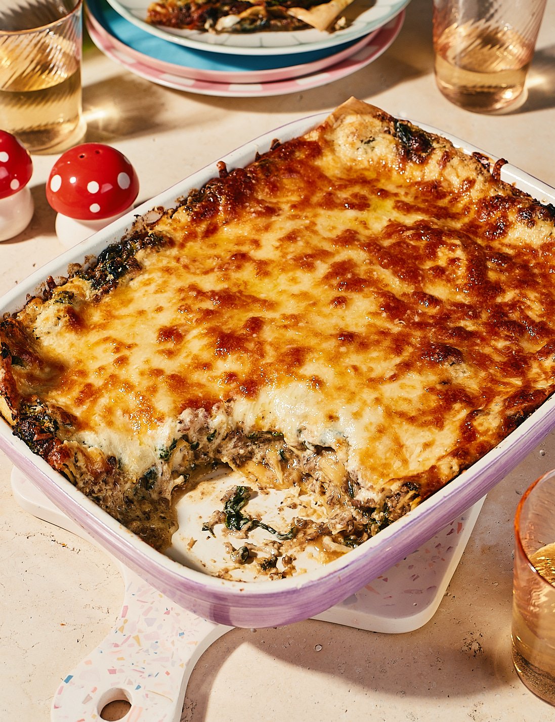 Baked lasagna in a rectangular dish with a golden cheese crust, partially served on a white and pastel-colored handle tray. Surrounding it are two glasses of sparkling drink, two red-and-white mushroom-shaped salt and pepper shakers, and a table with