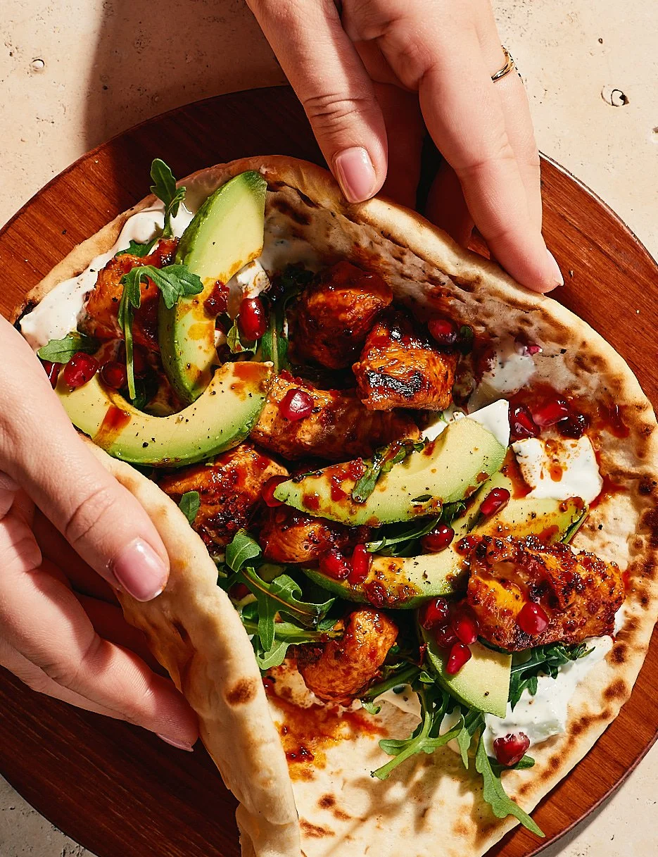 Person holding a pizza with chicken, avocado, arugula, pomegranate seeds, and sauce on a wooden plate.