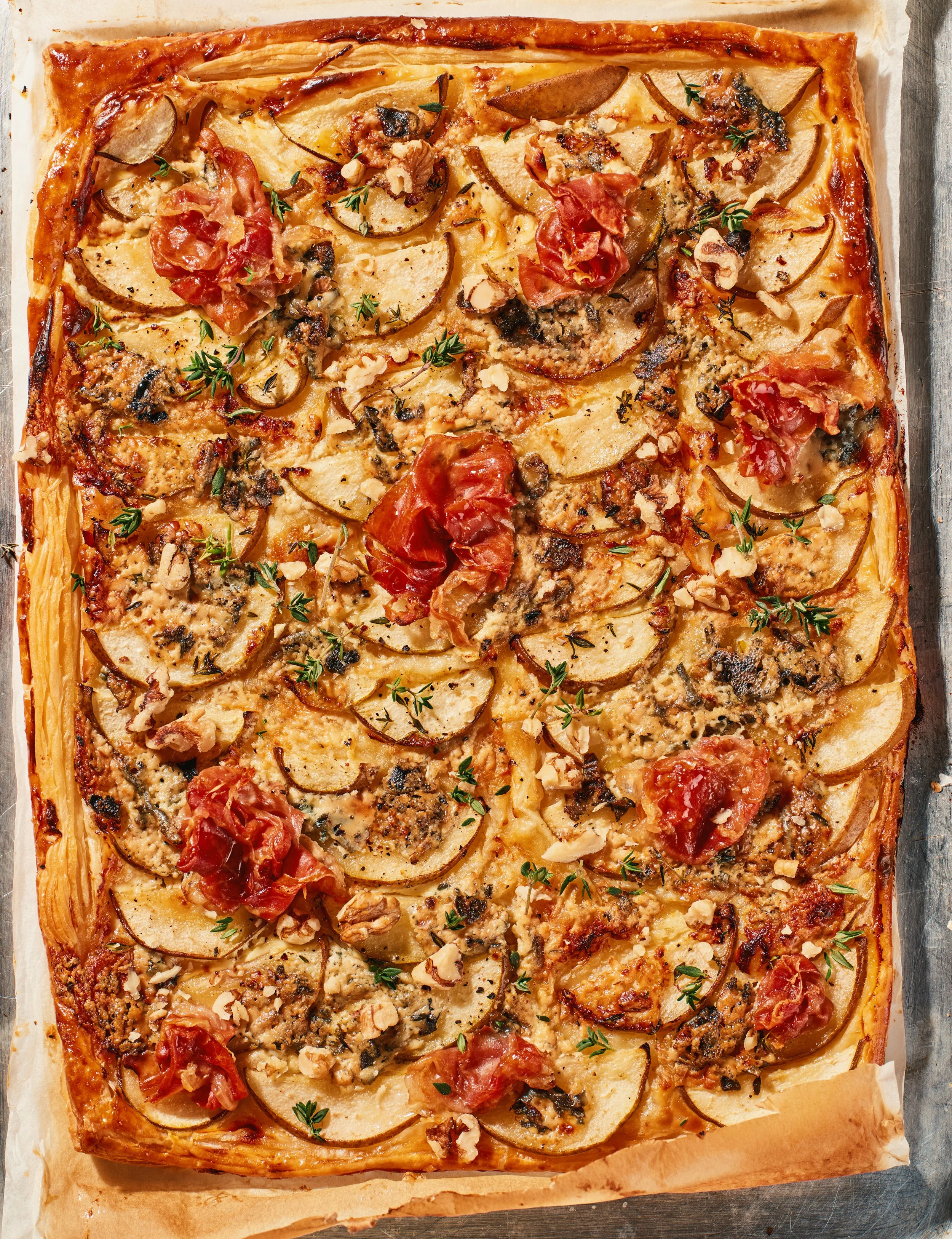 A rectangular vegetable tart with a golden-brown crust topped with sliced pears, cherry tomatoes, chopped walnuts, fresh thyme sprigs, and black pepper on a parchment paper-lined baking sheet.