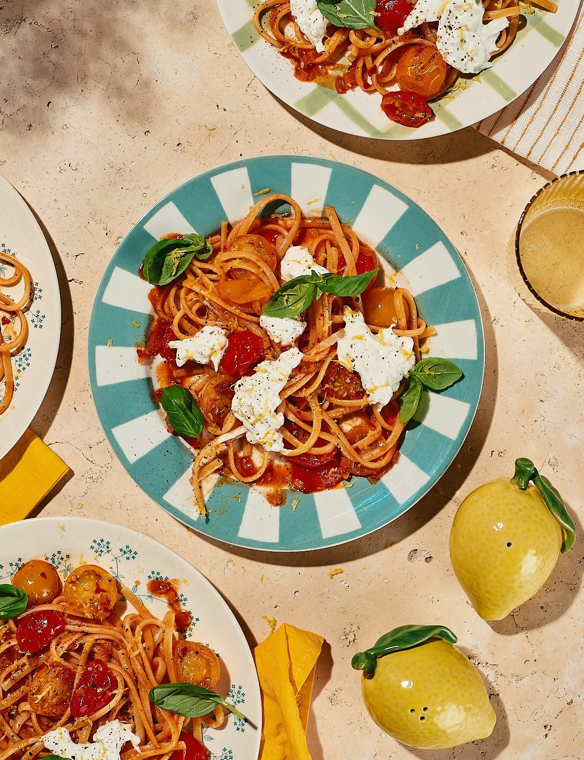 Plate of spaghetti with cherry tomatoes, fresh basil, and dollops of ricotta cheese, served on a colorful striped plate. There are additional plates of pasta, a glass of white wine, and ceramic lemon-shaped pitchers on a light-colored table.