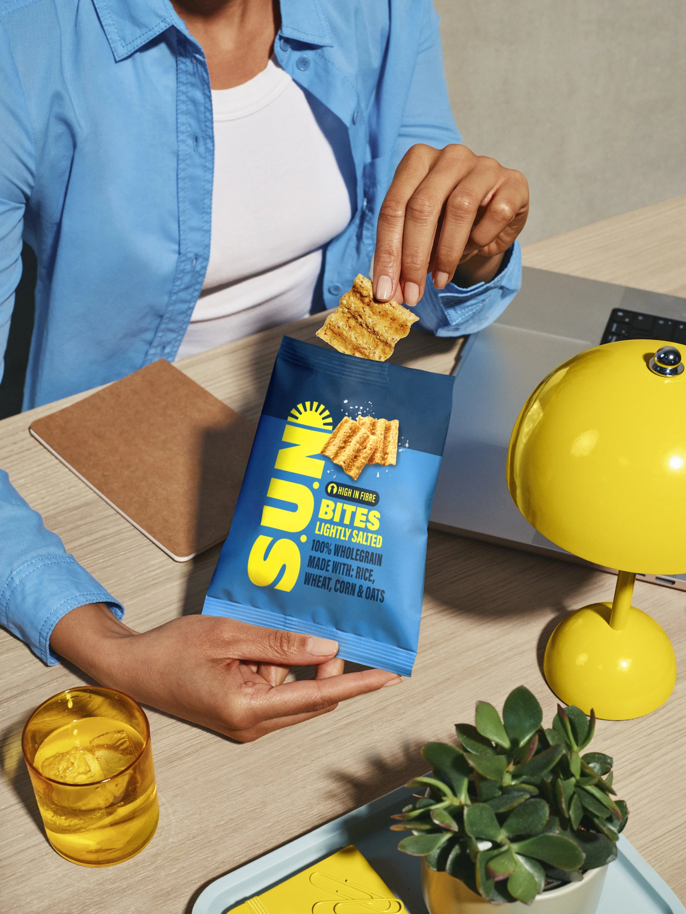 A person wearing a blue shirt and white T-shirt, holding a blue bag of sunflower snack bites, sitting at a desk with a yellow desk lamp, a plant, a glass of water with ice, and office supplies.