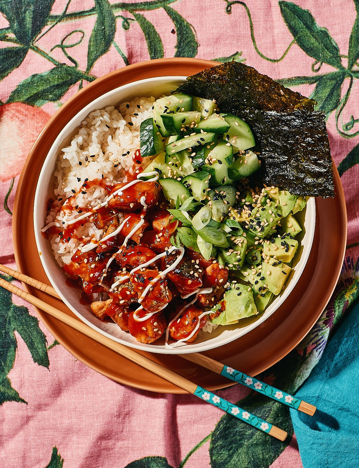 A bowl of rice with sesame seeds, topped with glazed chicken pieces garnished with white sauce, next to cucumber salad with sesame seeds, and diced avocado with sesame seeds, wrapped with a sheet of nori seaweed, all on a pink floral tablecloth with 