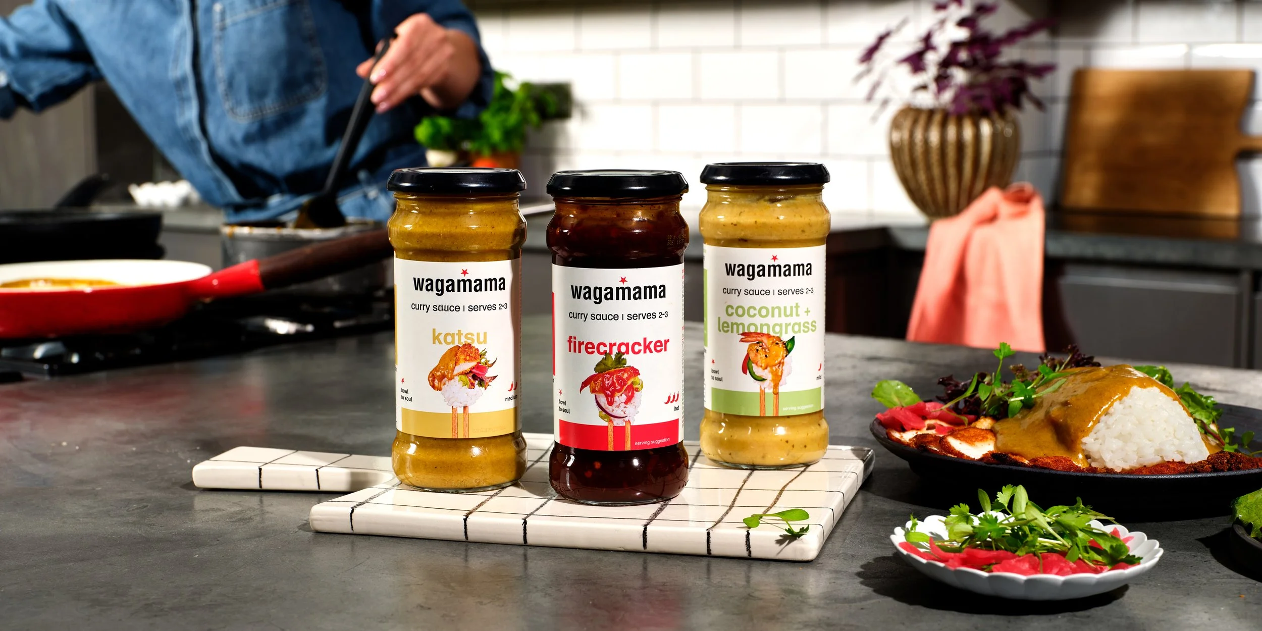Three jars of Wagamama curry sauces labeled "katsu," "firecracker," and "coconut + lemongrass" on a white and black striped cutting board on a kitchen countertop, with a plate of rice and curry, green herbs, and a person cooking in the background.