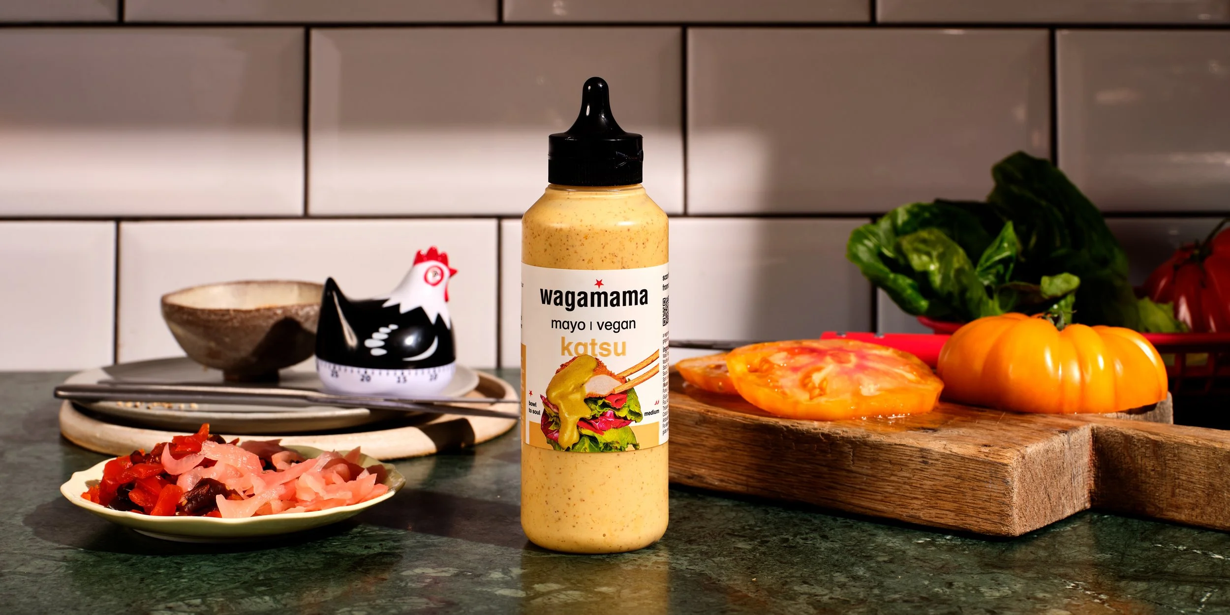 Bottle of wagamama vegan katsu mayo on a kitchen counter, with vegetables and ingredients surrounding it, including a small bowl, a plate of pickled onions, a wooden cutting board with tomato, greens, and a red basket with peppers.