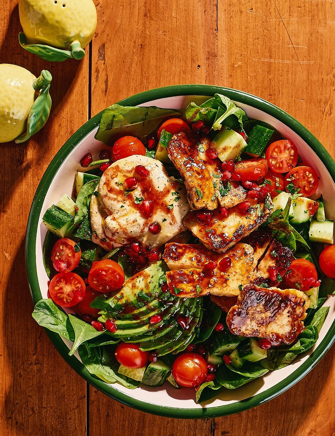 Fresh salad with cherry tomatoes, cucumber, avocado, grilled pieces of chicken, poached egg, pomegranate seeds, and greens on a plate. Two yellow ceramic lemon-shaped salt shakers are on a wooden table.