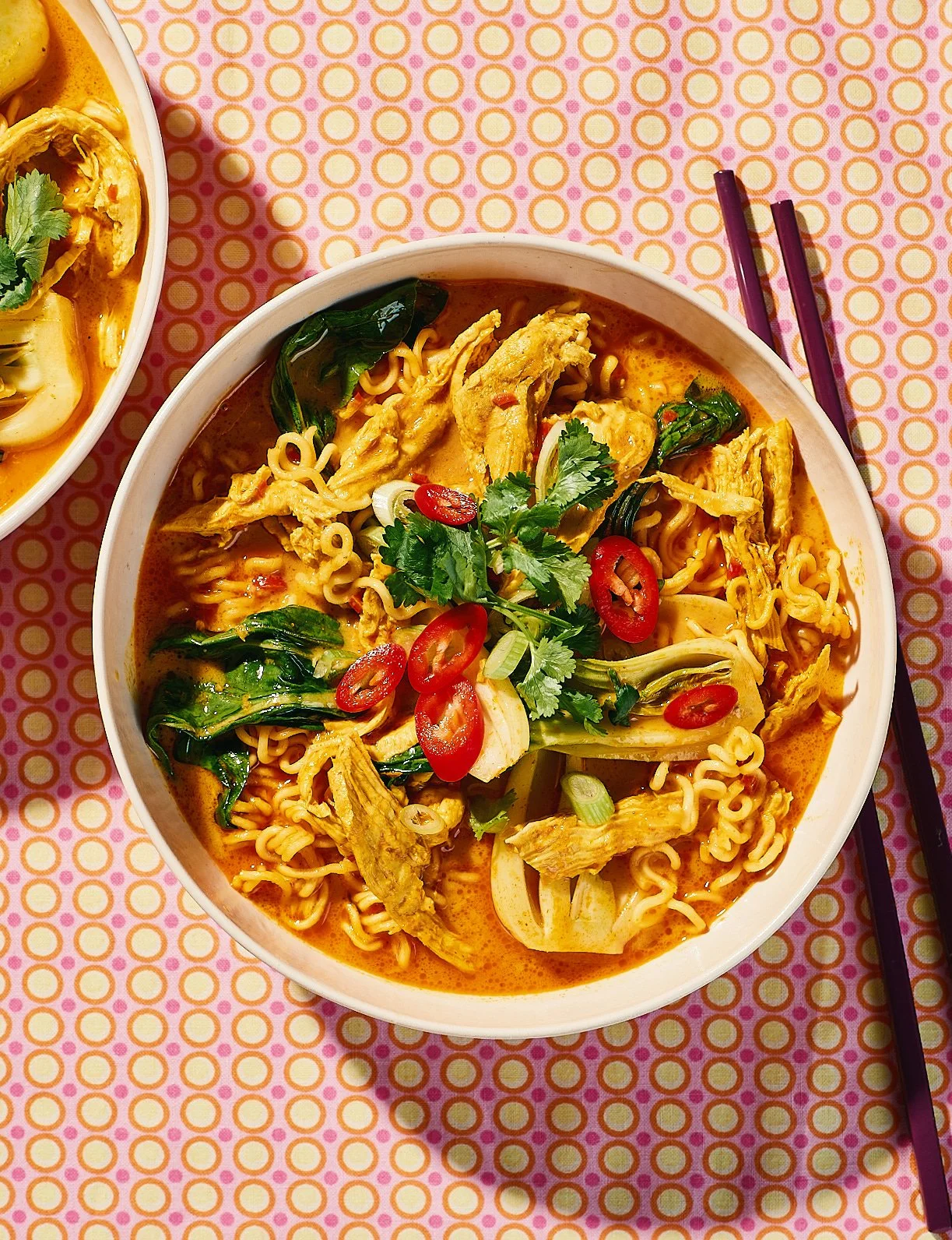 A bowl of spicy ramen noodle soup with chicken, bok choy, sliced red chili peppers, and garnished with cilantro and green onions, on a pink patterned tablecloth with purple chopsticks.
