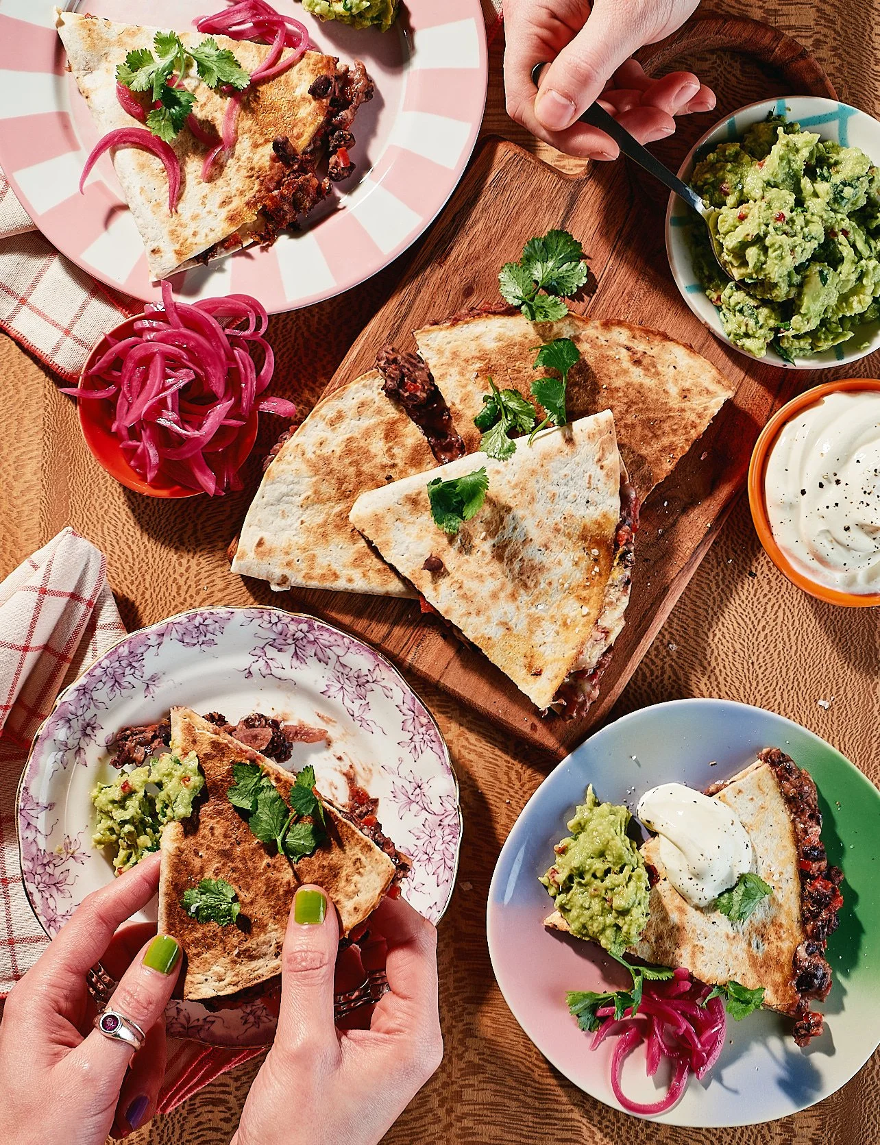A table of Mexican dishes including quesadillas, burritos, guacamole, pickled red onions, and sour cream, with a hand holding a piece of quesadilla in the foreground.