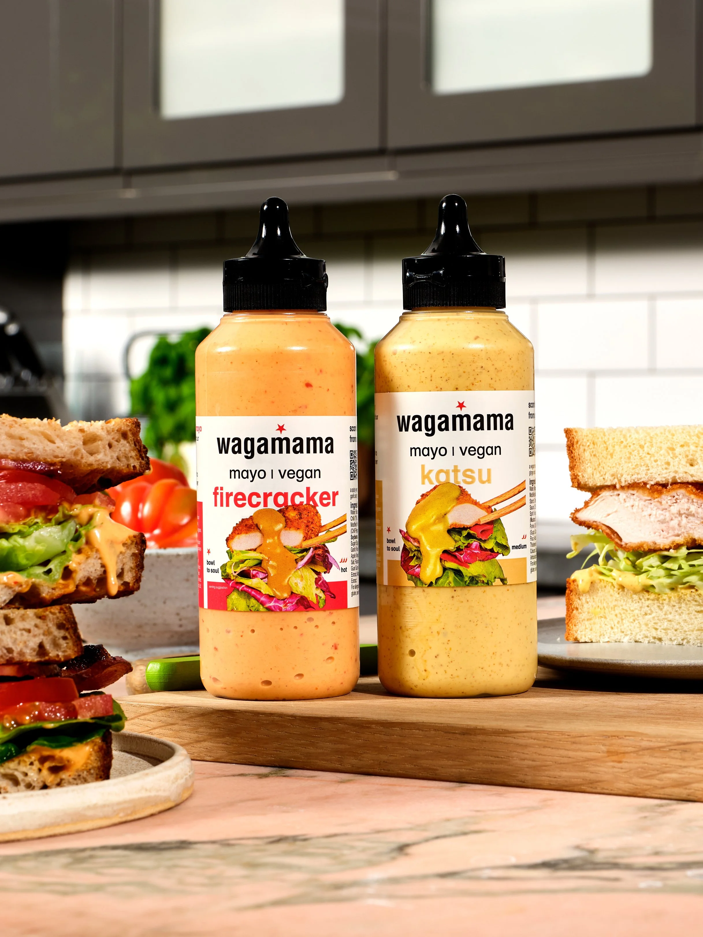 Two bottles of wagamama vegan sauces, firecracker and katsu, placed on a wooden cutting board with sandwiches and ingredients nearby in a kitchen setting.