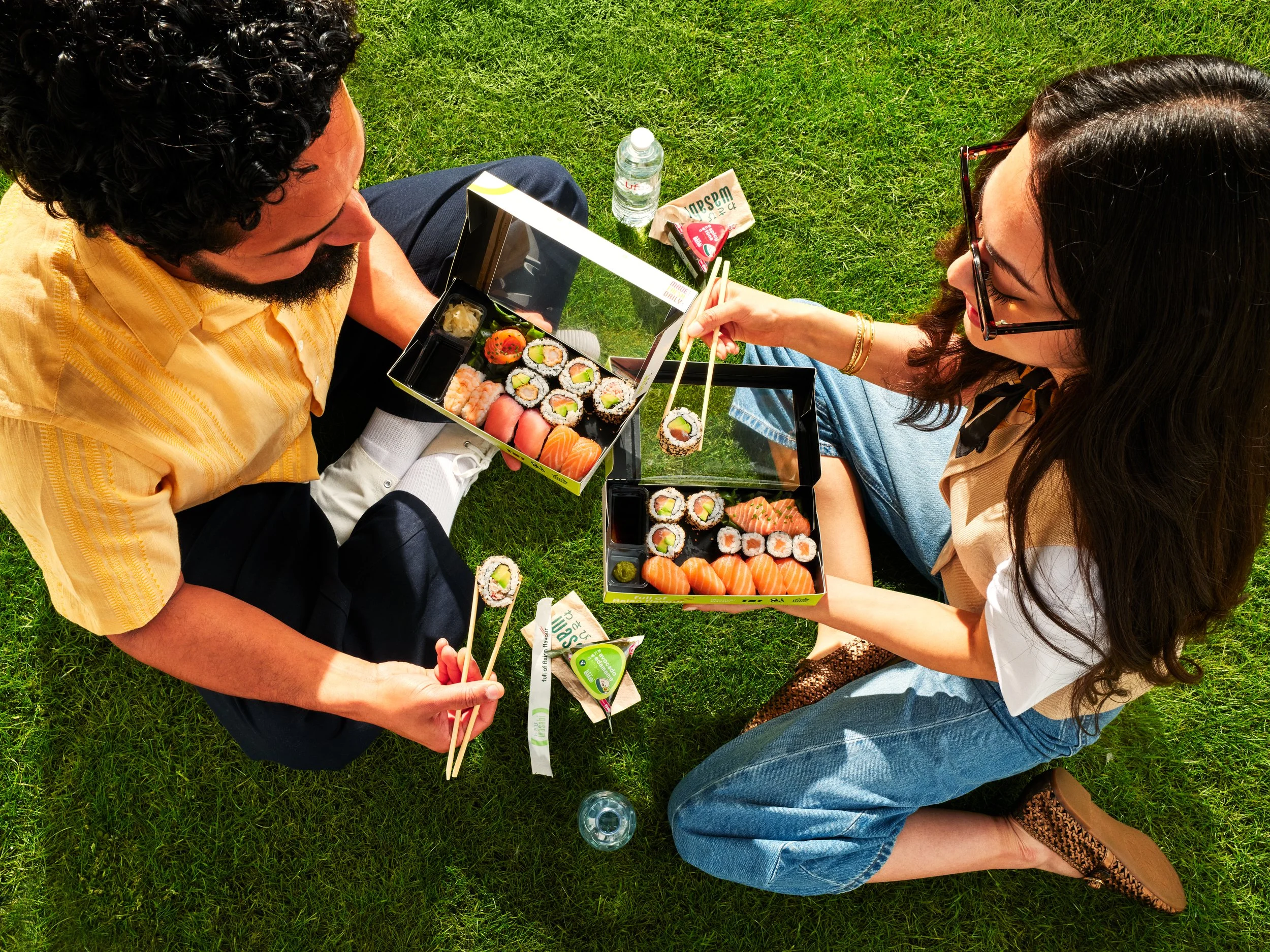 Two people sitting on grass enjoying Wasabi sushi from a black takeout box, with a water bottle and snack items nearby.