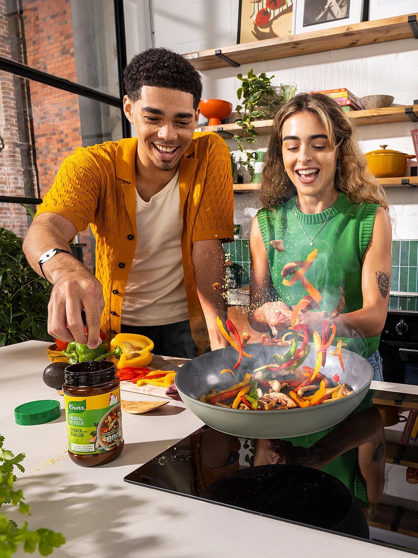 Two people cooking together in a kitchen with shelves, plants, and artwork on the wall. They are adding vegetables to a frying pan, with one person smiling and the other focused. There are sliced vegetables and a jar of chicken bouillon on the counte
