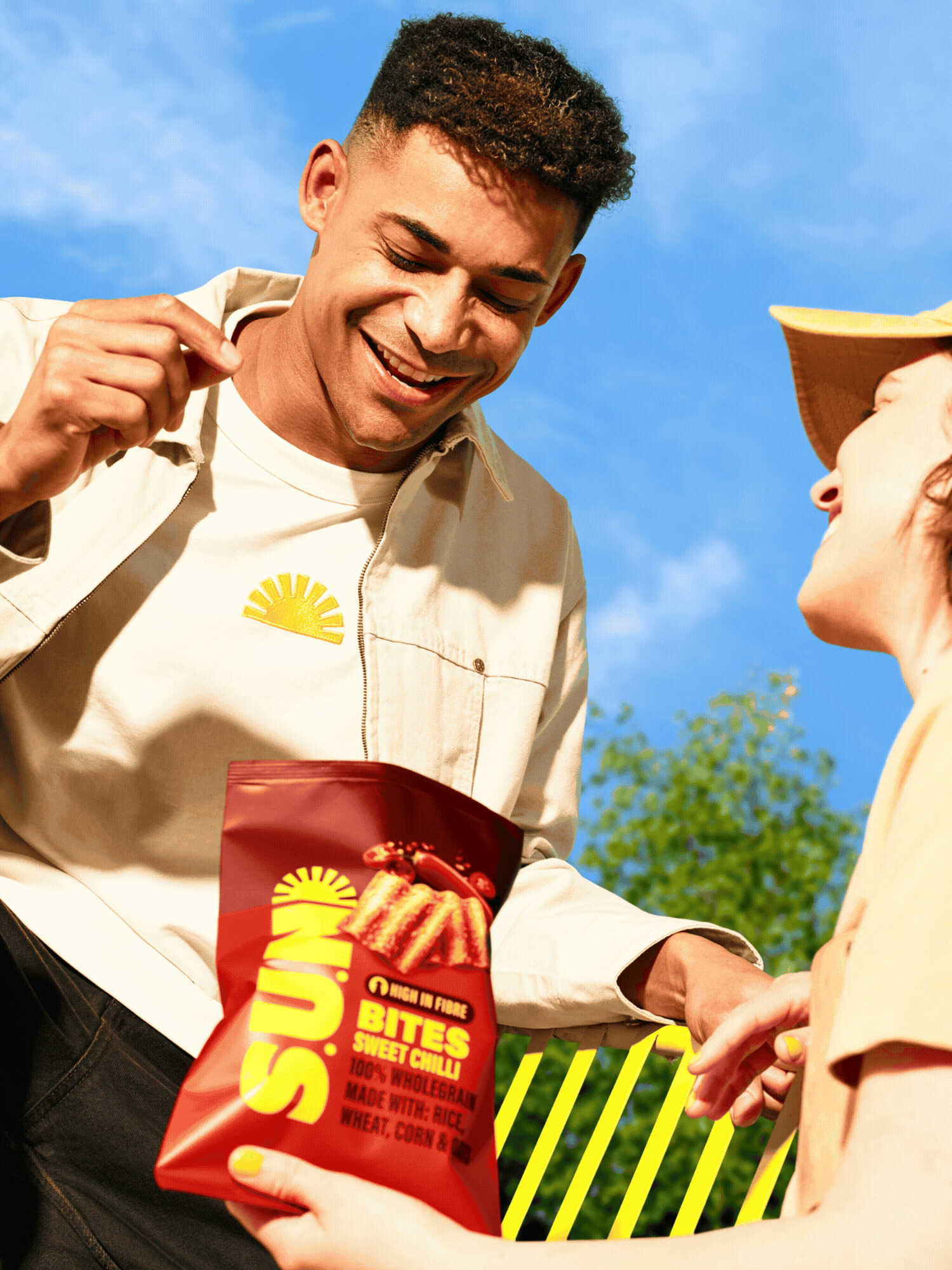 A man with short dark curly hair smiling and looking down is holding a bag of spicy sweet chili flavored snack chips, while a woman with a wide-brimmed hat and glasses is handing him the snack outdoors with a blue sky and trees in the background.