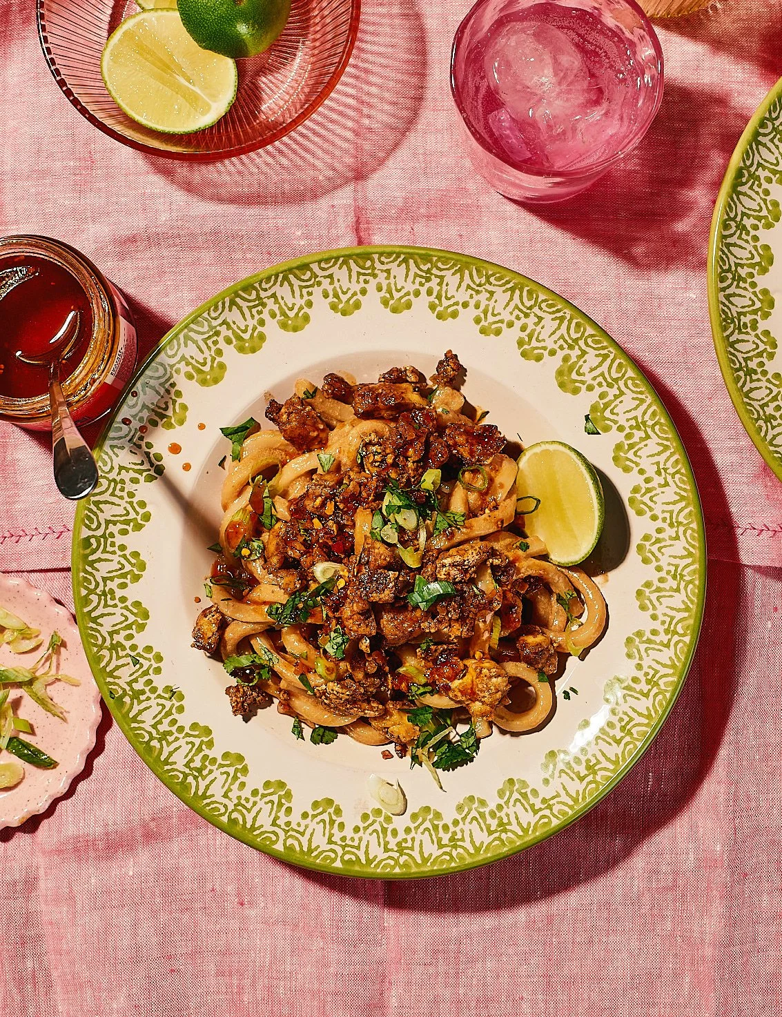 Plate of beef and pasta with chopped green onions and herbs, served with lime, on a green and white patterned plate, surrounded by small bowls of lime, lime halves, and a jar of sauce, on a pink tablecloth.