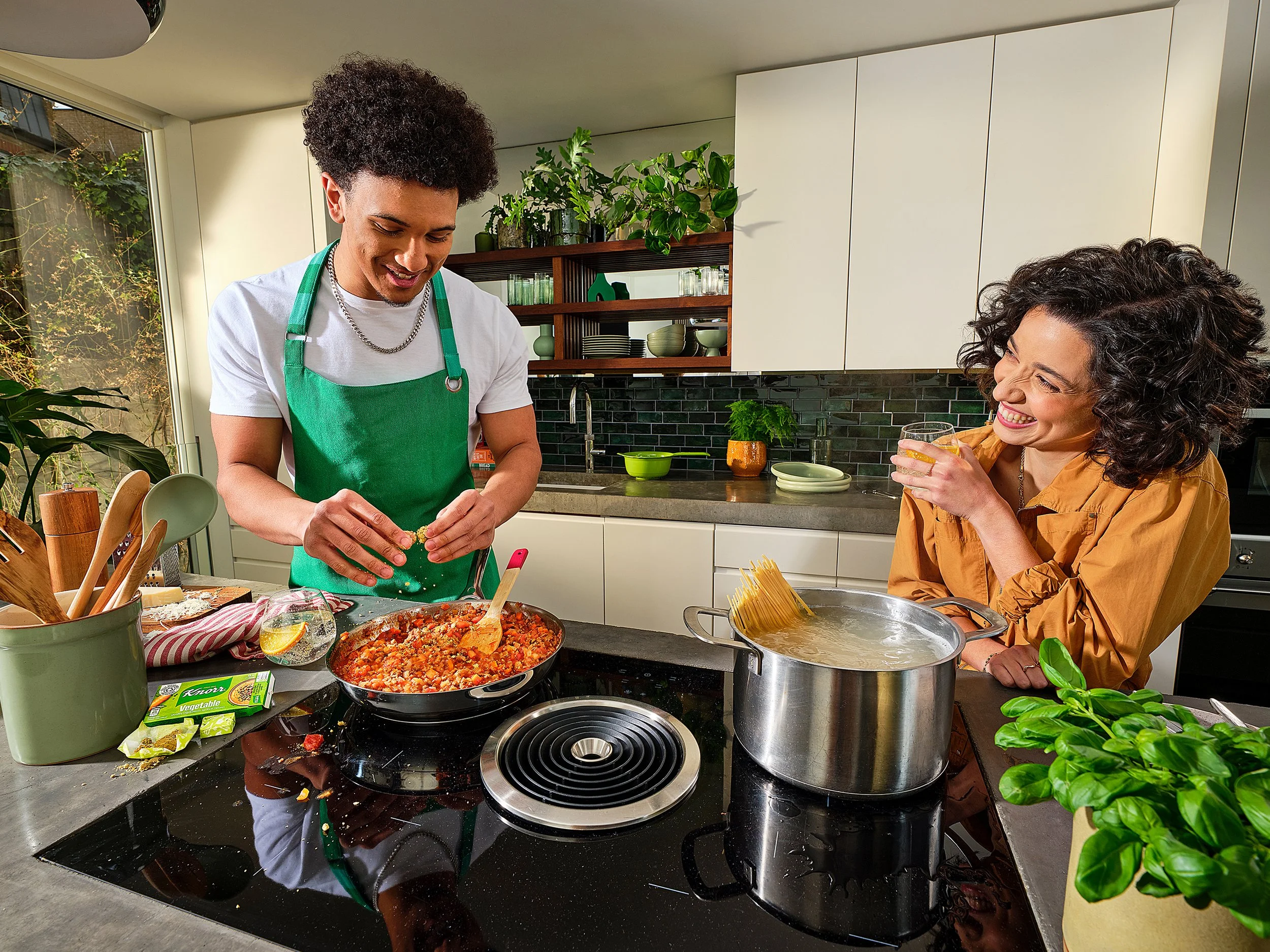 Two women are in a modern kitchen, one is preparing food with a pan of cooked spaghetti with tomato sauce on the countertop, while the other is sitting, smiling, and drinking a beverage. The kitchen has bright lighting, green plants, and open shelves