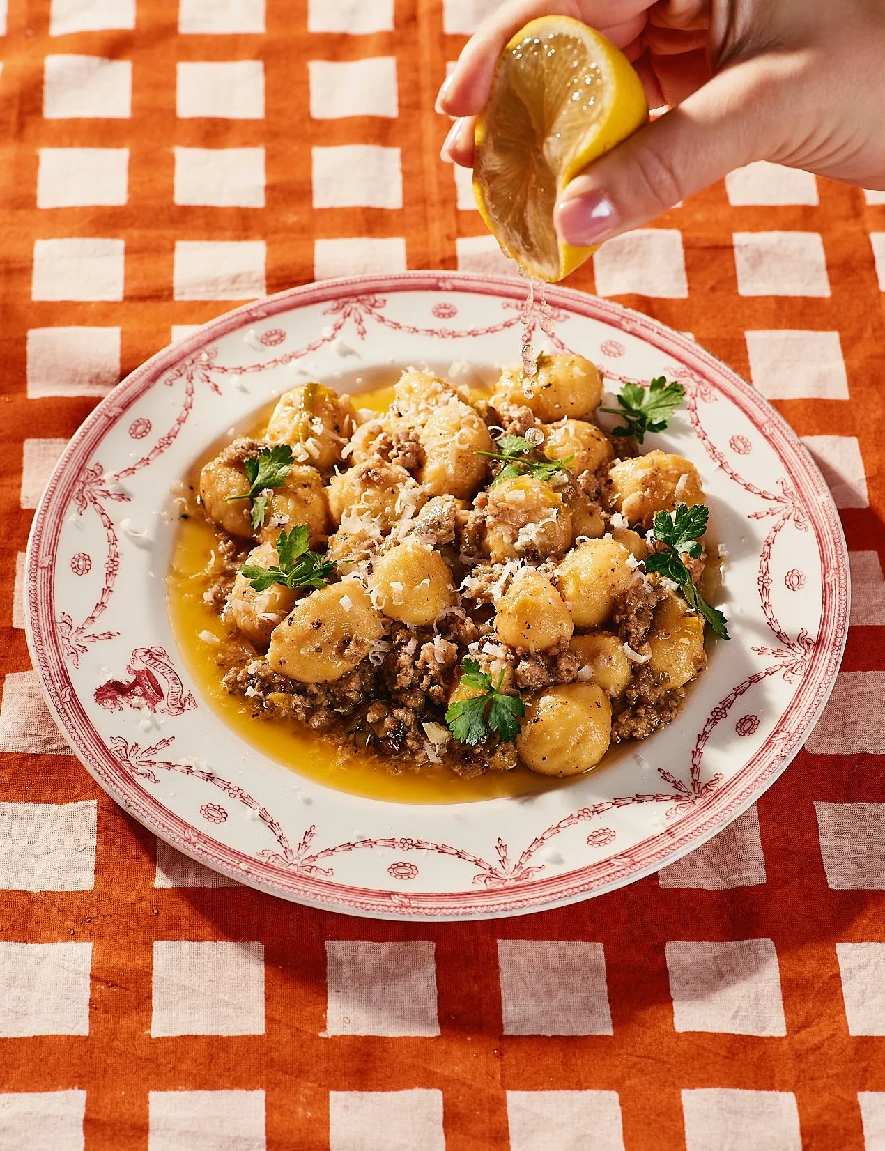 A plate of gnocchi with ground meat, garnished with parsley, and topped with grated cheese, on a red and white checkered tablecloth. A hand is squeezing lemon over the dish.