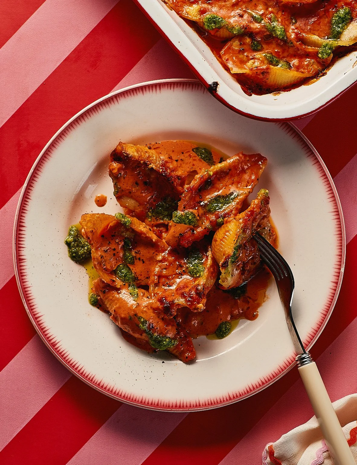 Plate of cooked chicken with sauce and herbs, served on a white plate, with a fork. In the background, a dish of baked chicken with vegetables in a red and white baking dish. Both dishes are on a red and pink striped tablecloth.
