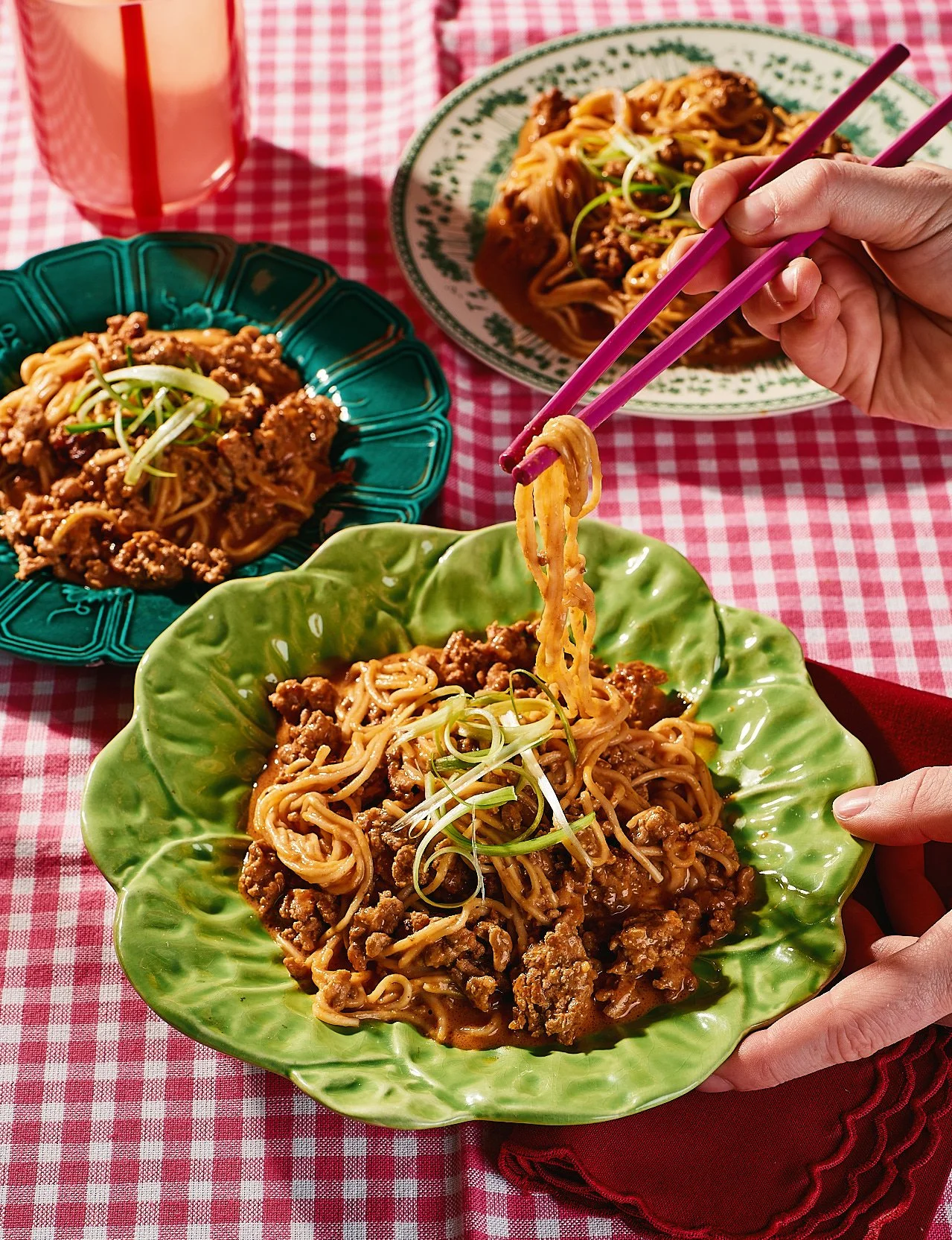 A colorful meal of Asian stir-fried noodles with minced meat and green onions served on a green, leaf-shaped plate, with additional plates of similar dish in the background and a drink in a glass with a red straw on a red and white checkered tableclo