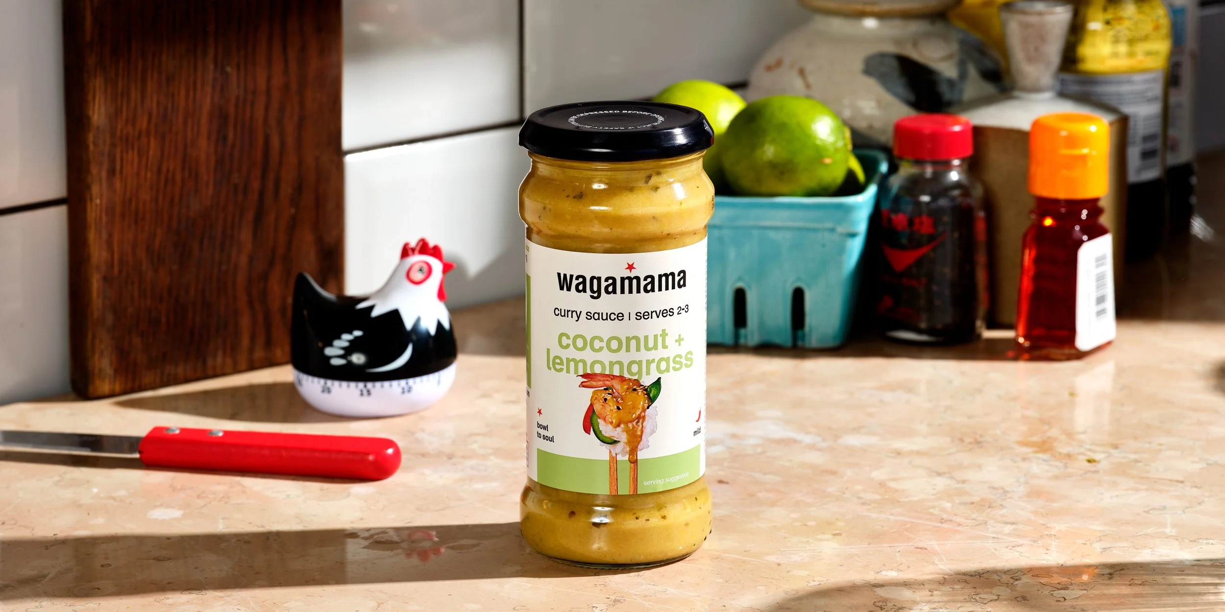 A jar of Wagamama coconut and lemongrass curry sauce on a kitchen counter with various jars, apples, and a decorative chicken figurine in the background.