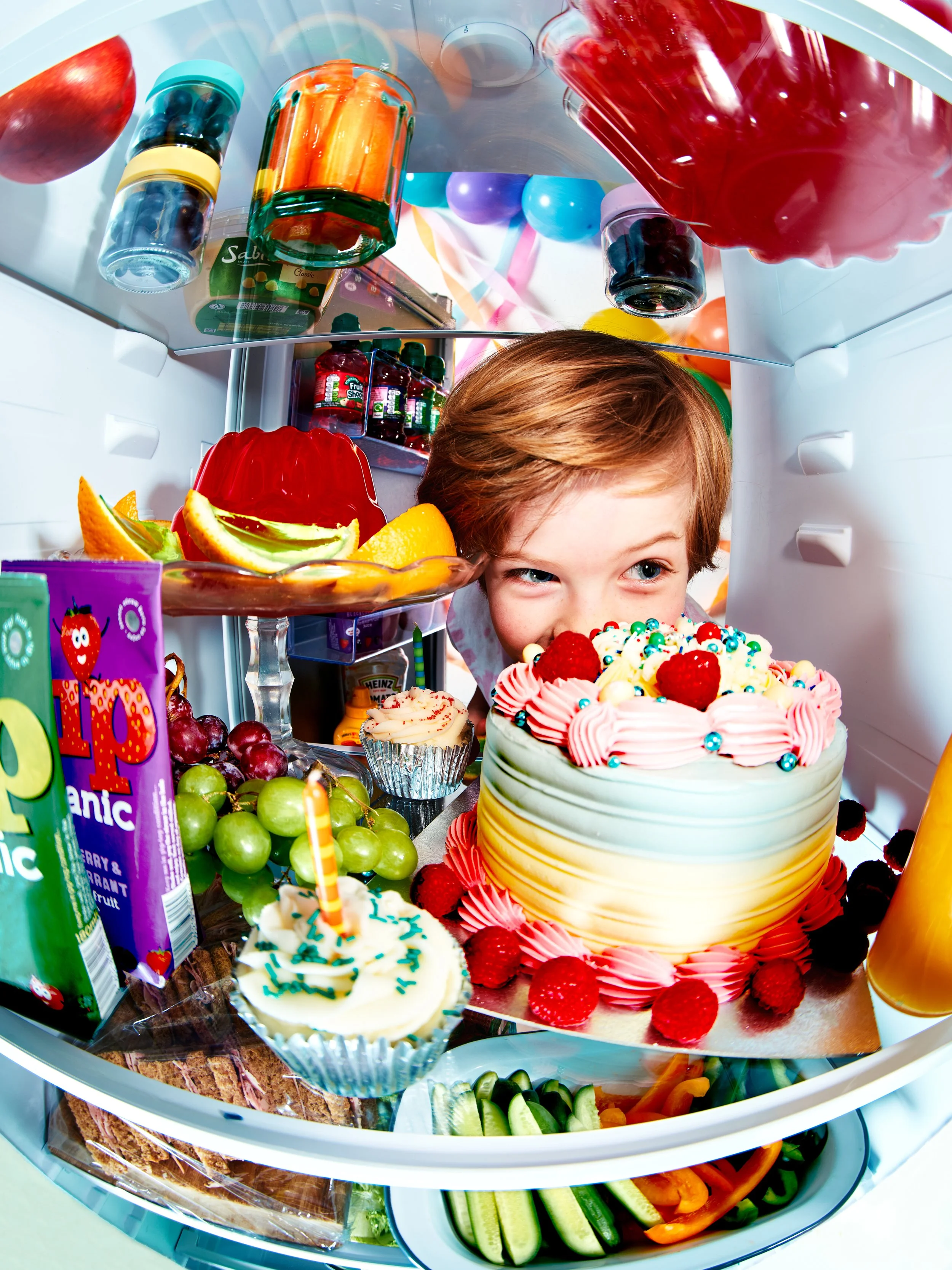 Inside a refrigerator filled with various colorful foods, desserts, and beverages, including a decorated cake, cupcakes, fruit, candy, and bottles of liquids.
