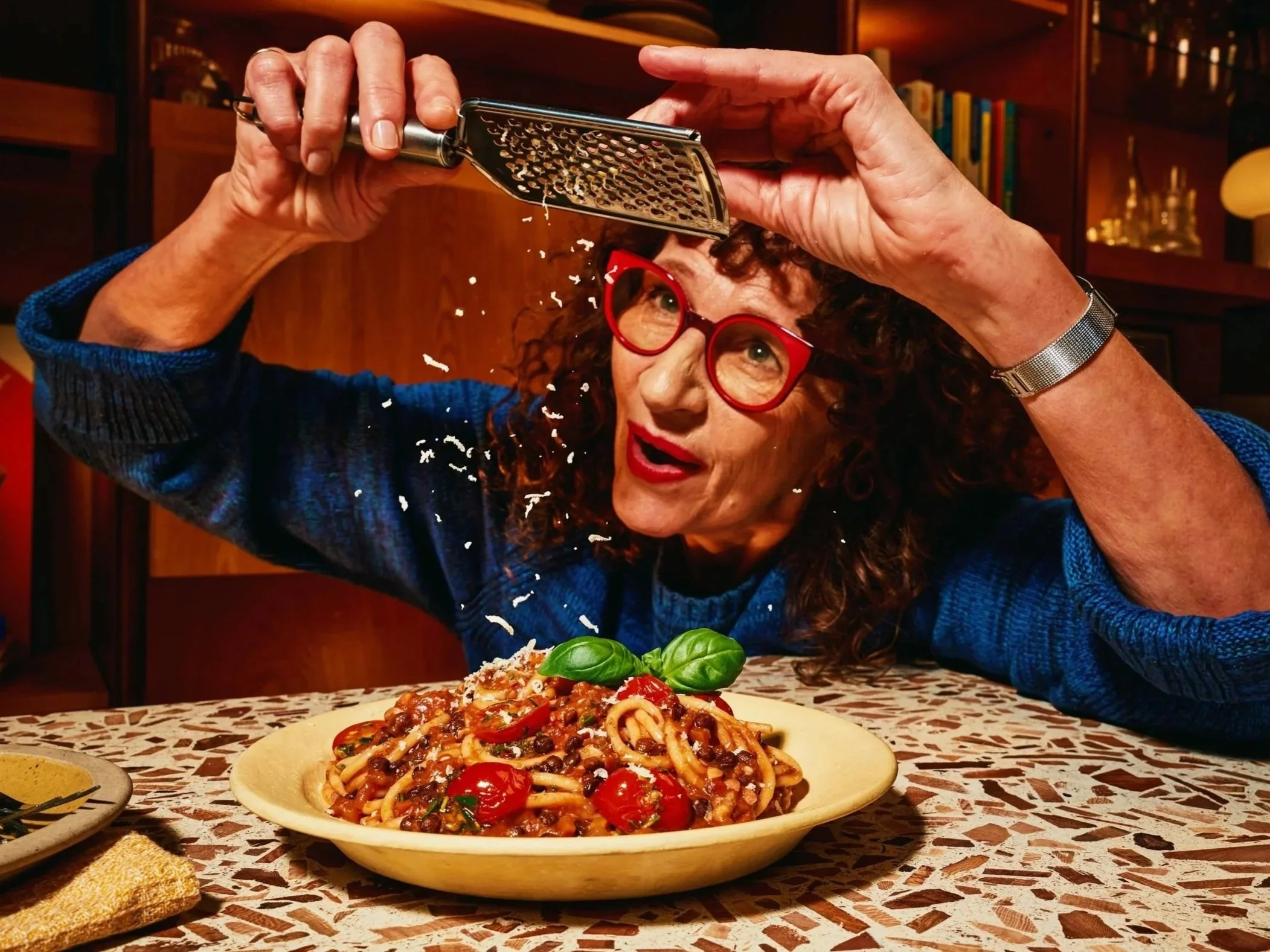 A woman with curly hair and red glasses is grating cheese over a plate of spaghetti with tomato sauce, black beans, and cherry tomatoes, sitting at a table with a patterned tablecloth.