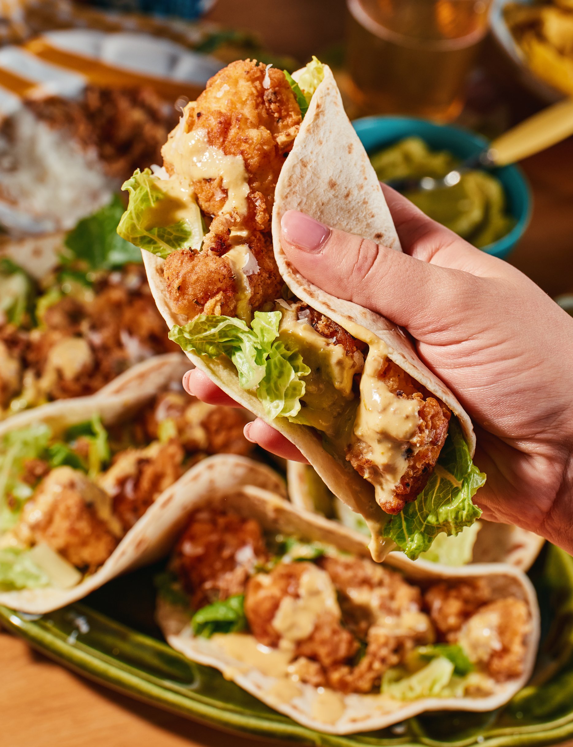 A hand holding a taco filled with fried chicken, lettuce, and cheese. In the background, there are more tacos and bowls of garnishes or sides on a platter.