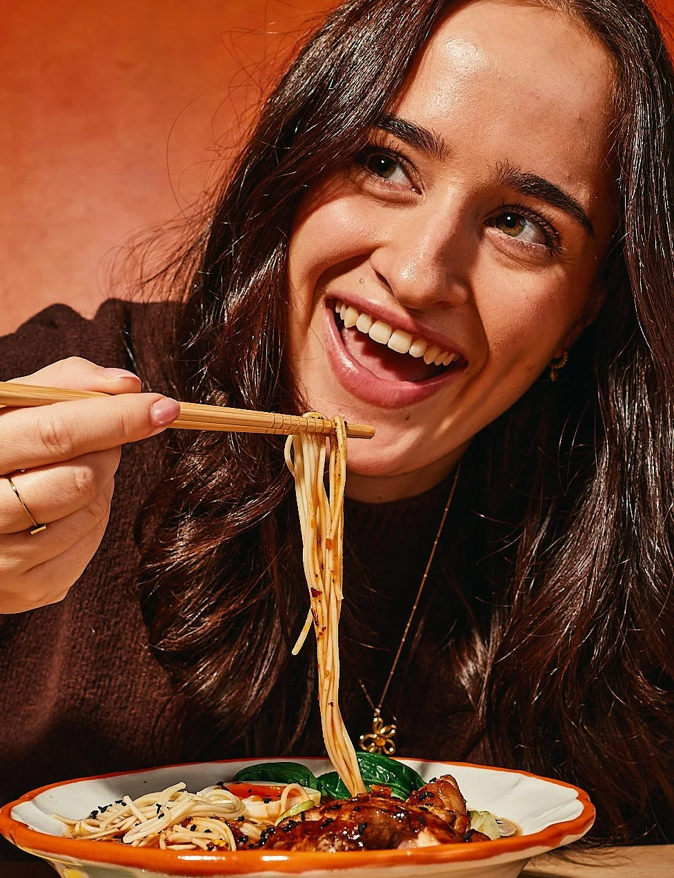 A woman with dark hair smiling while eating noodles with chopsticks at a bowl of Asian cuisine.