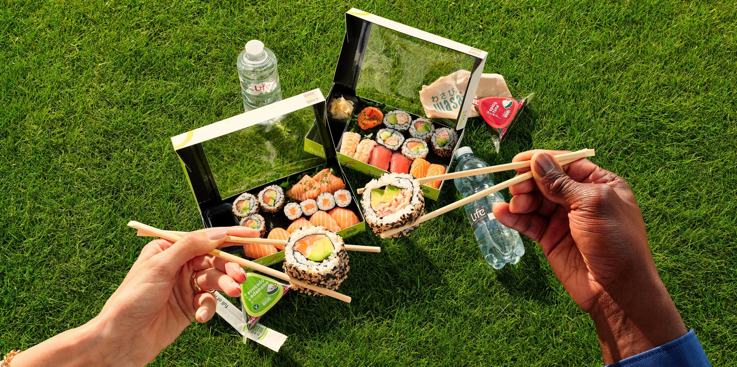 Hands holding chopsticks picking up a roll of Wasabi sushi with a green avocado filling, with a sushi box, water bottles, and snacks on green grass.