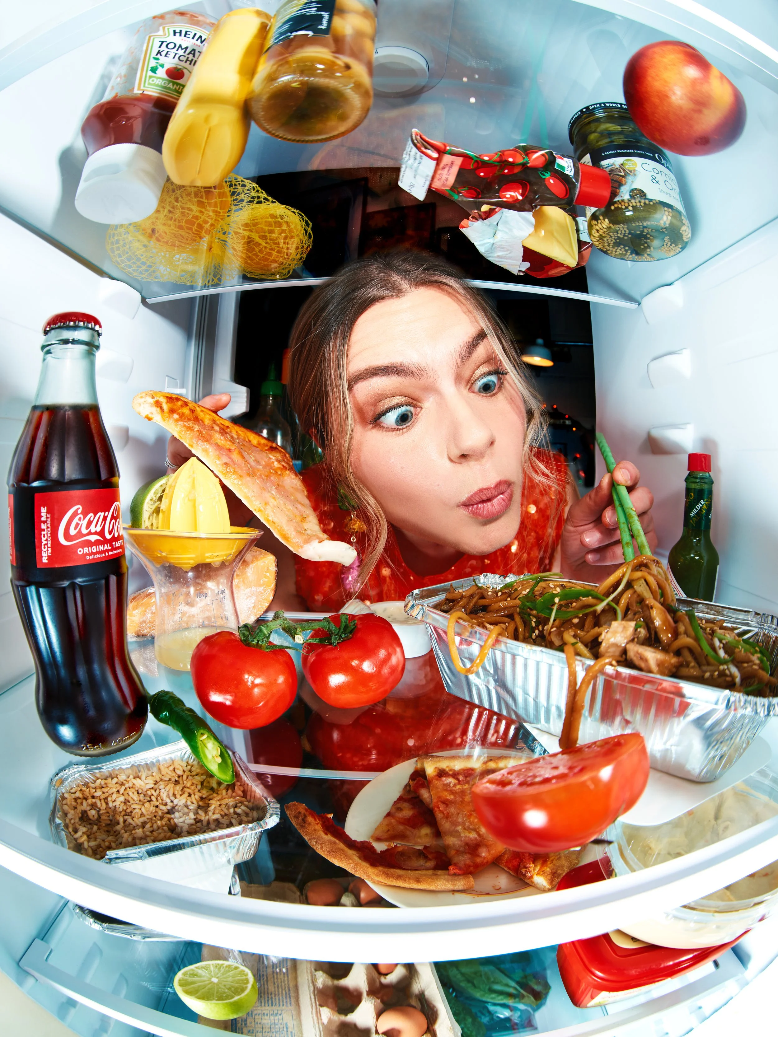 Open refrigerator door with various food items including tomatoes, pizza slices, a bowl of noodles, a bottle of Coca-Cola, a lemon half, an apple, ketchup, mustard, and mustard seeds.