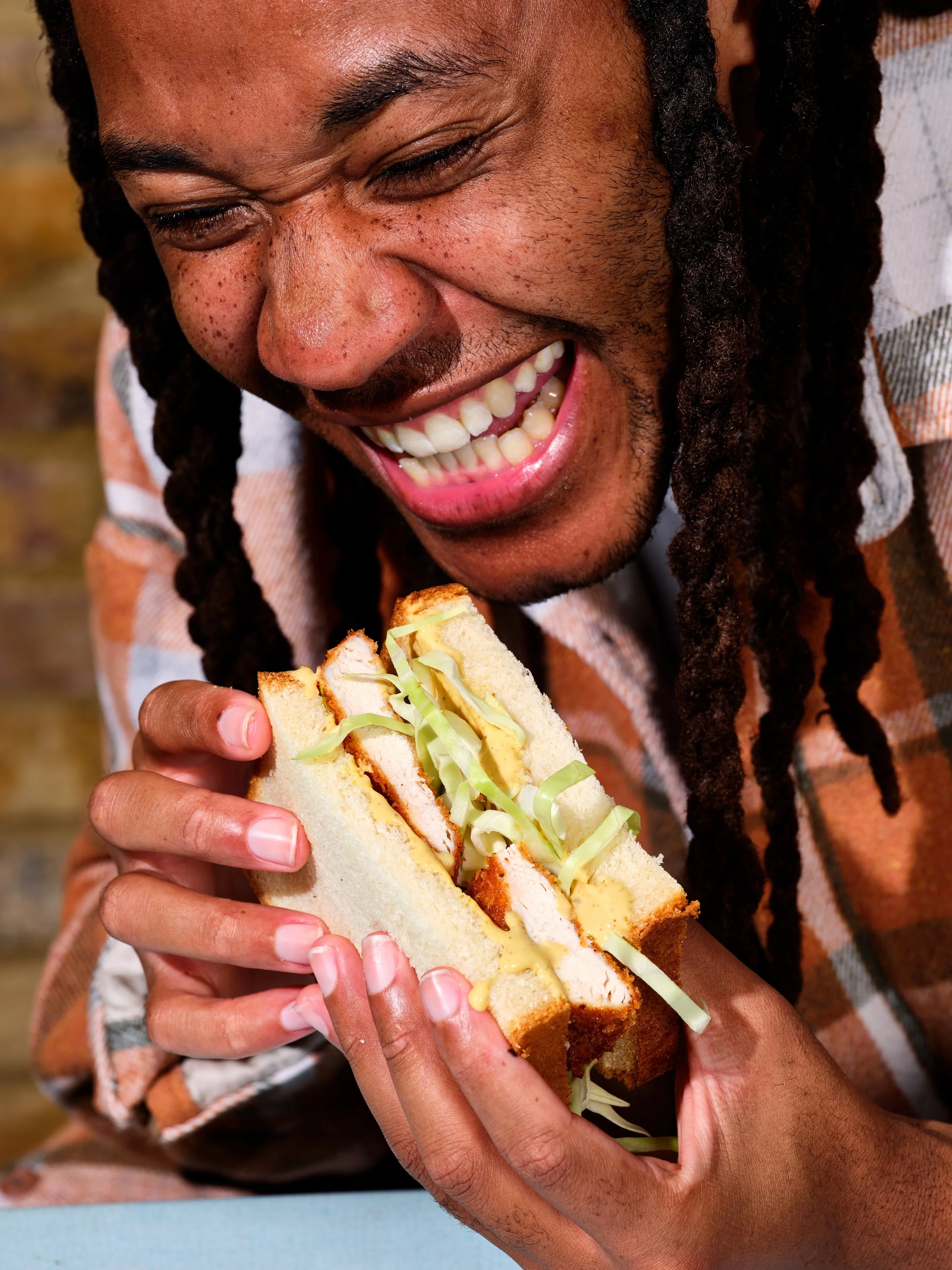 A person with long dreadlocks smiling and holding a partially eaten sandwich with fried chicken, lettuce, and mustard.