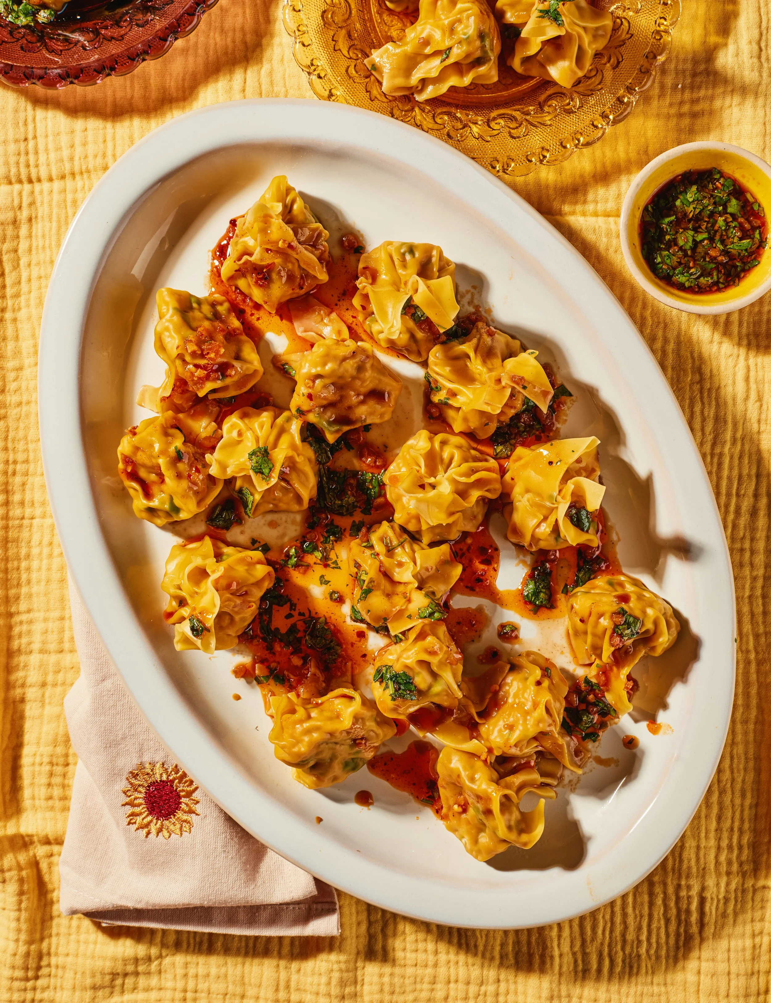 A white oval platter filled with cooked dumplings topped with sauce and herbs, placed on a yellow tablecloth with a sunflower design on a napkin, alongside a small bowl of dipping sauce and another dish of dumplings in a glass bowl.