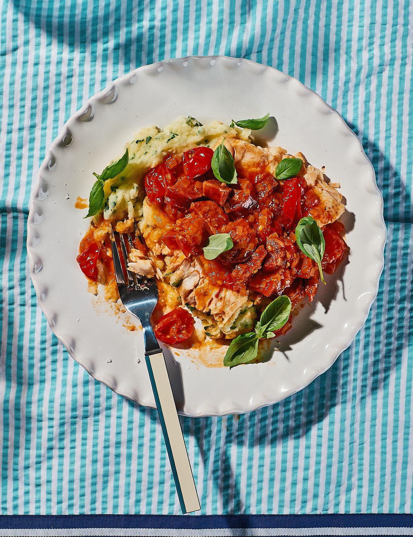 A plate of fish topped with tomato sauce, garnished with basil leaves, served with mashed potatoes on a white scalloped plate, placed on a blue and teal striped tablecloth.