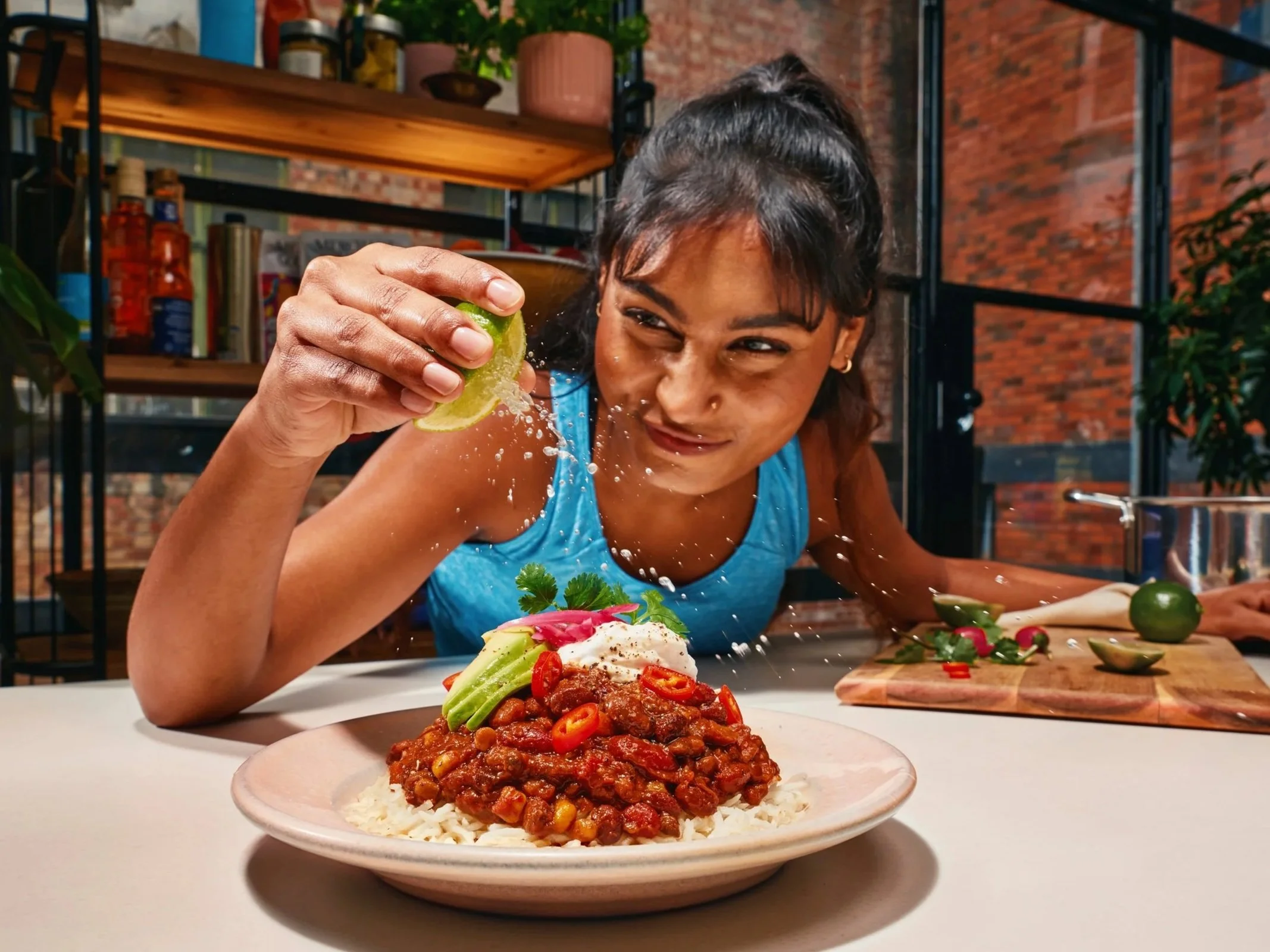 A woman with dark hair in a ponytail squinting as she squeezes lime over a plate of chili served over rice, with fresh vegetables and herbs on a dining table.