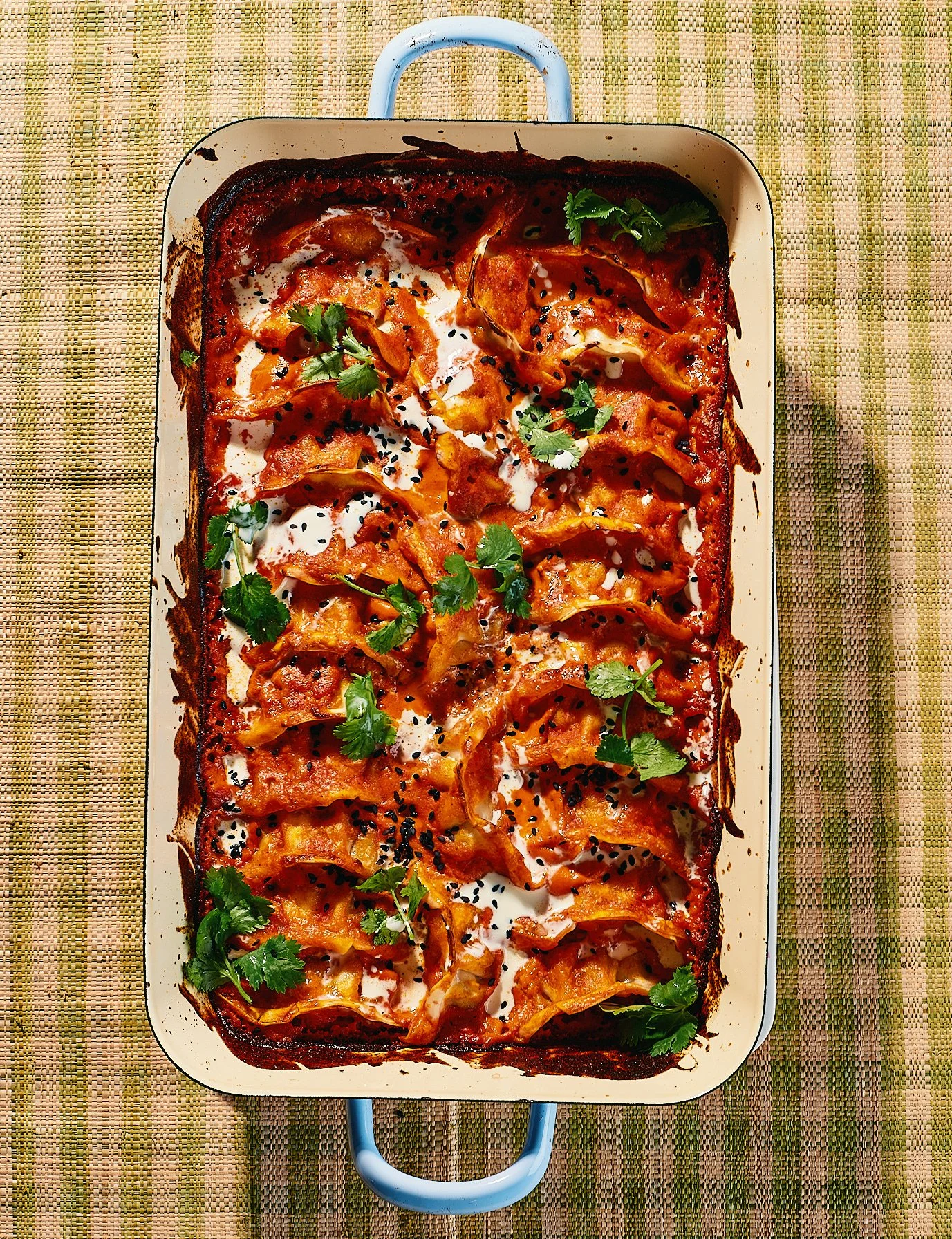 A baked casserole dish topped with melted cheese, sliced vegetables, cilantro, and black sesame seeds.