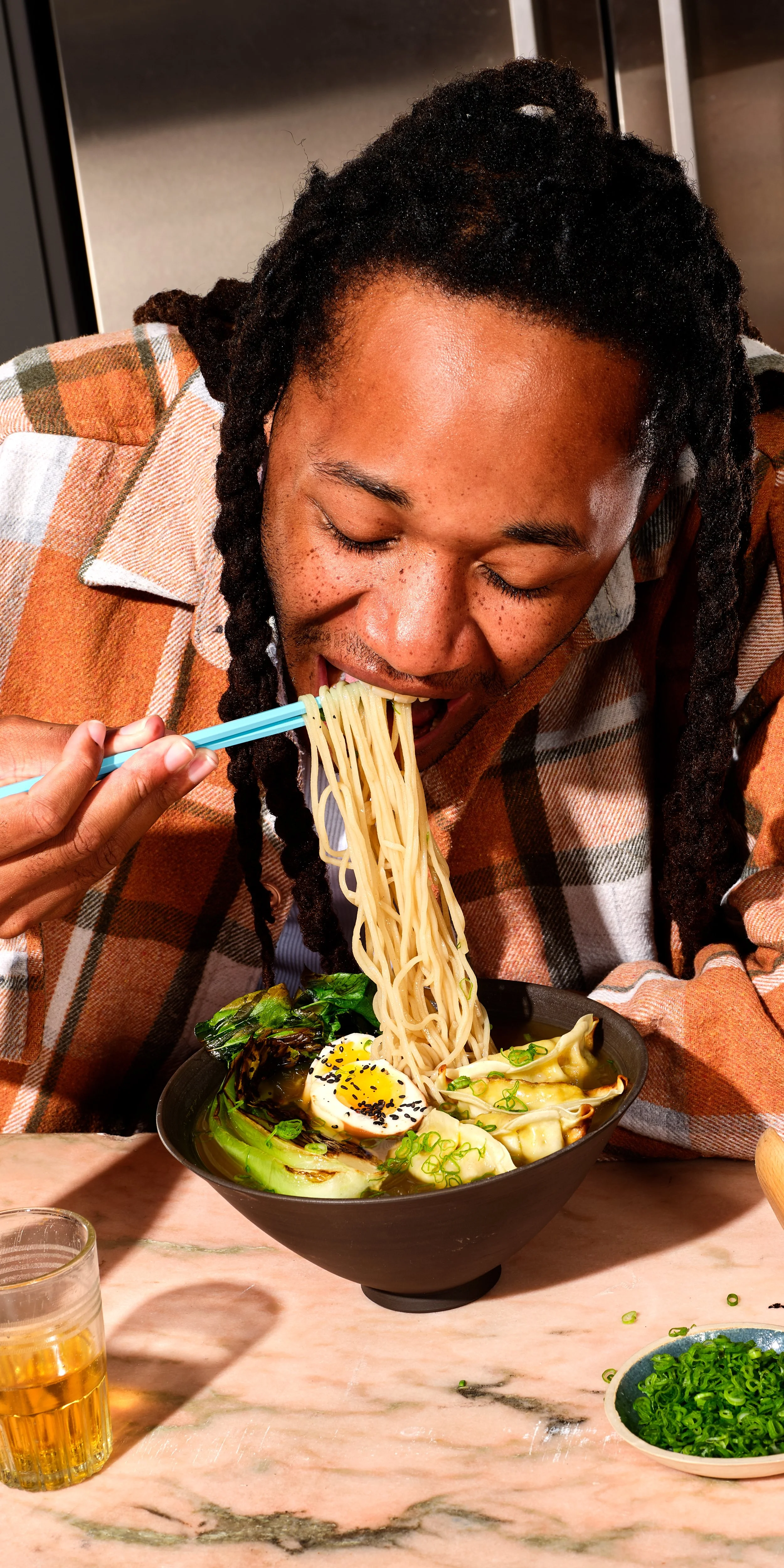 A person with dark hair styled in dreadlocks, wearing an orange checkered shirt, is eating noodles from a black bowl with a pair of blue chopsticks. The bowl contains ramen with a boiled egg, green onions, and leafy greens. There is a glass of beer a