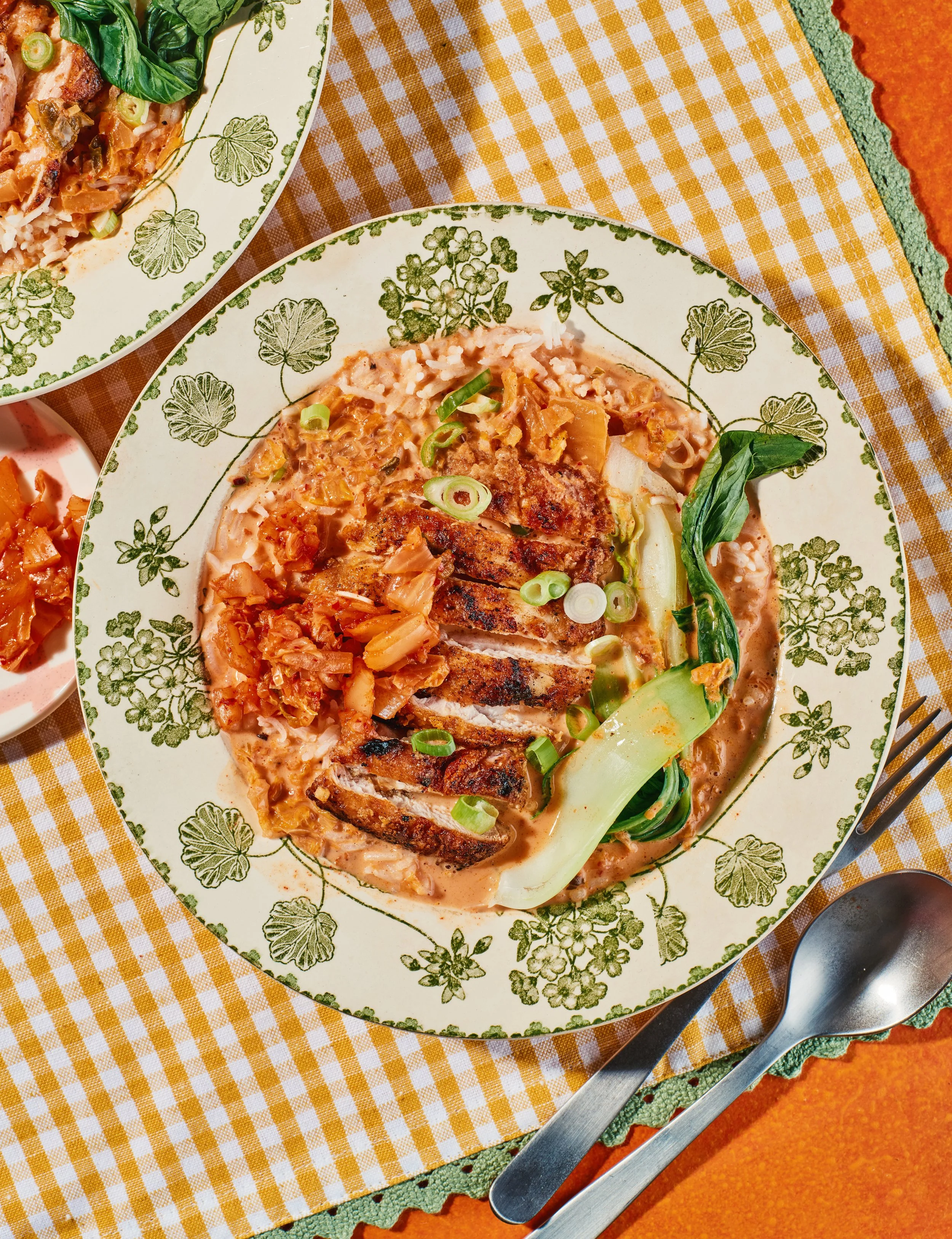 A plate of chicken fried steak with gravy, served with side dishes including kimchi, chopped green onions, and leafy greens, on a decorative plate with green floral patterns, placed on a yellow checkered tablecloth.