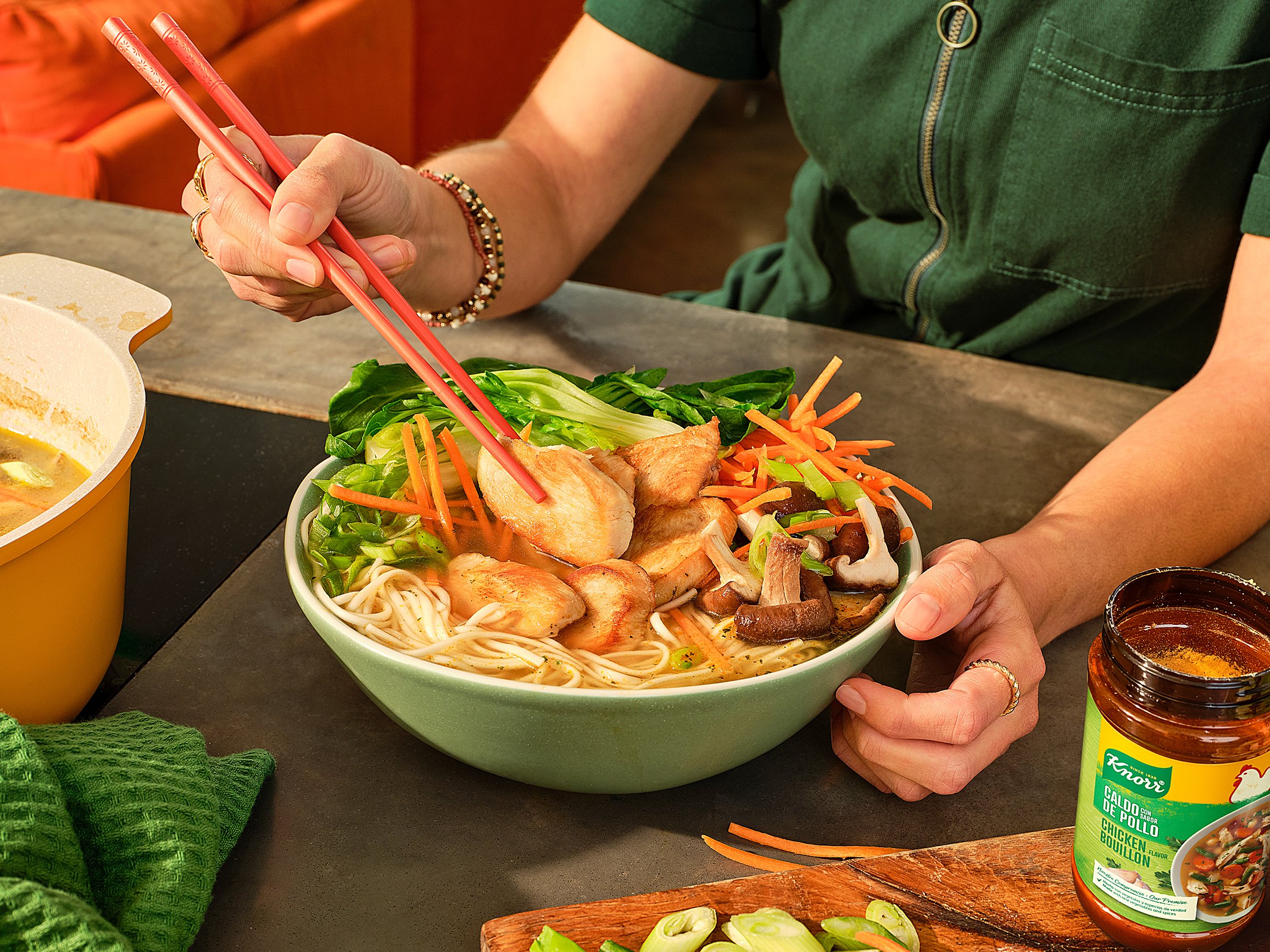 Person using red chopsticks to eat from a large bowl of Asian noodle soup with vegetables and chicken.