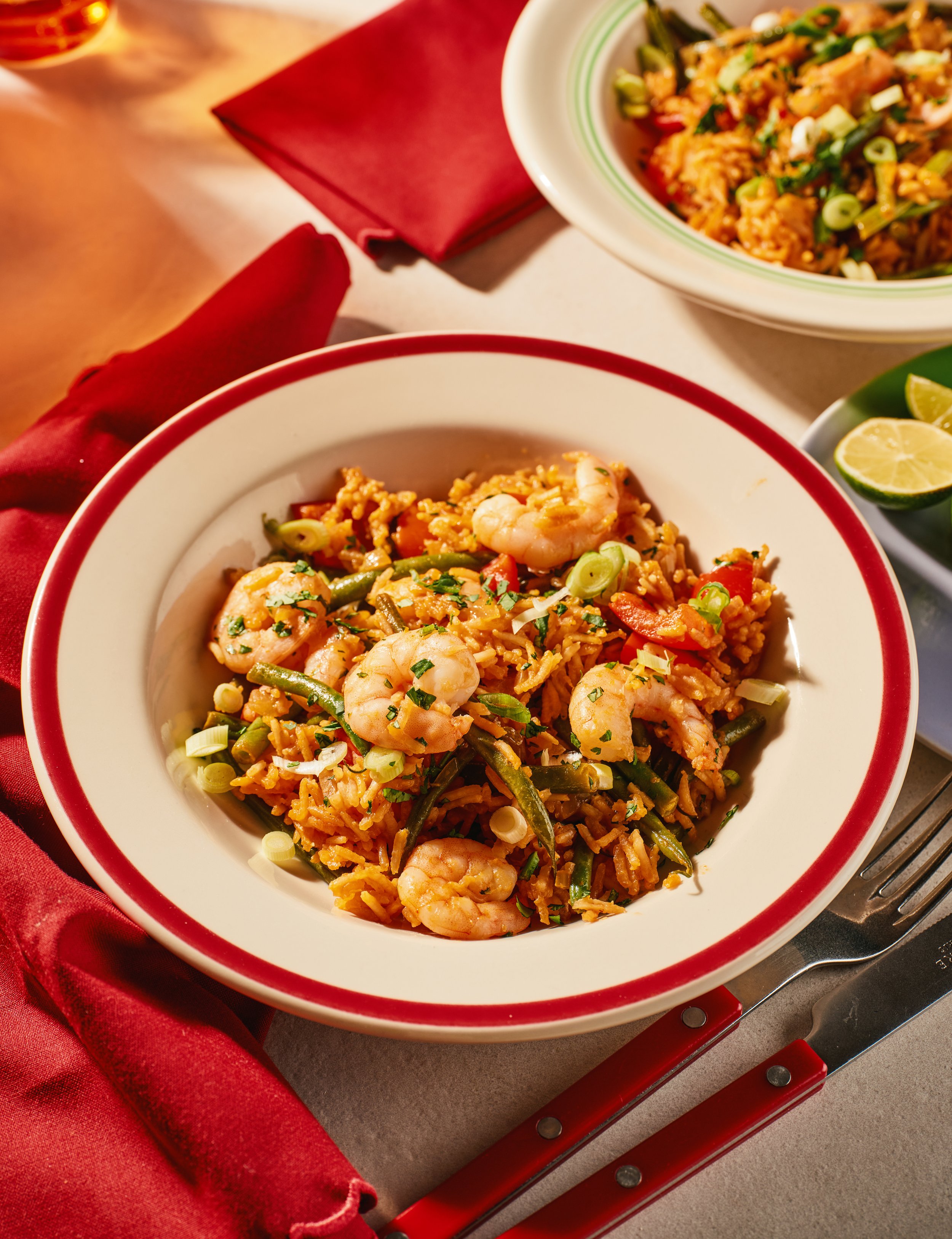A bowl of shrimp fried rice garnished with chopped green onions, set on a table with red napkins and lime wedges.